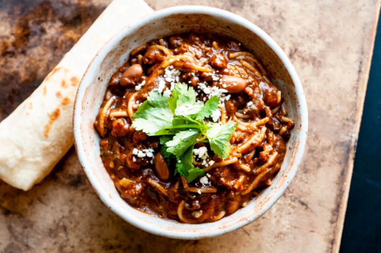 A bowl of chili with noodles, beans, and crumbled cheese, topped with cilantro and served with a flour tortilla.