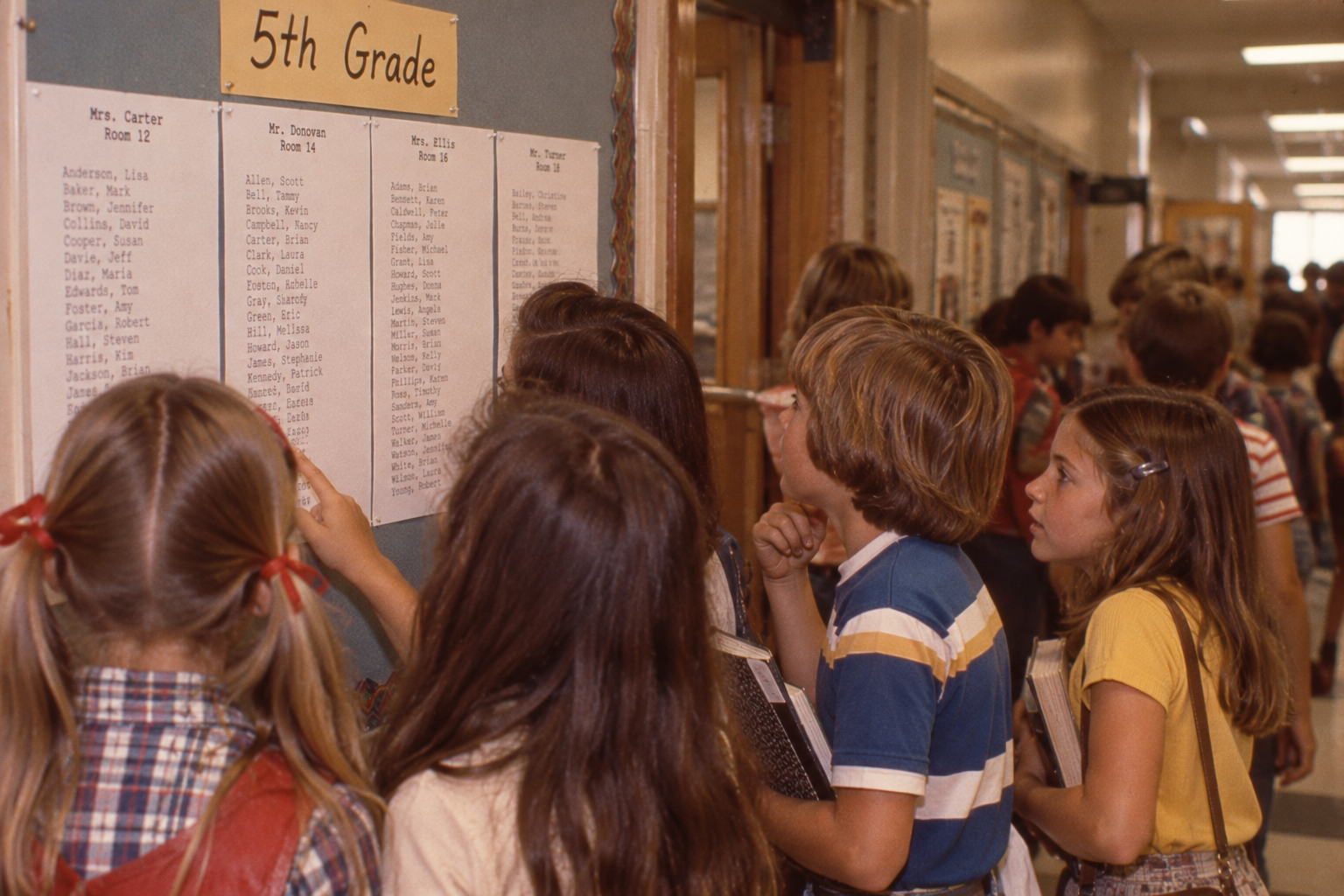 Students stand in a school hallway looking at a posted 5th grade class list.