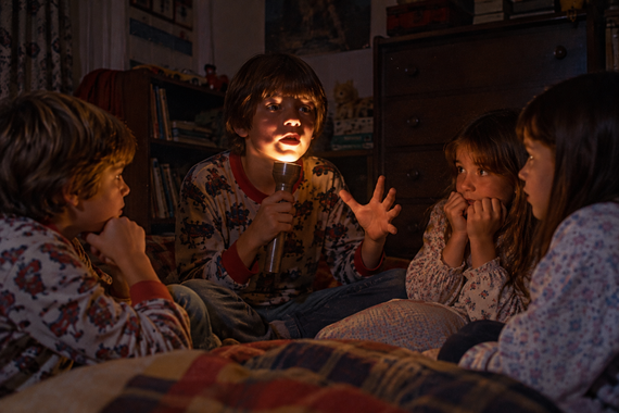 Four children sit in a circle in a dark room, one holding a flashlight up to their face to tell a story during a power outage.