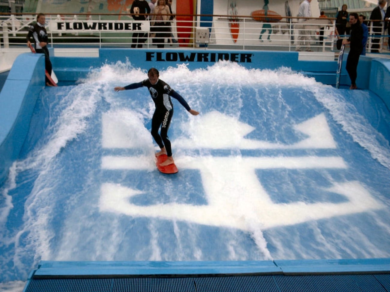 A person in a wetsuit stands on a surfboard on a FlowRider wave simulator, a popular cruise ship amenity found on the deck.