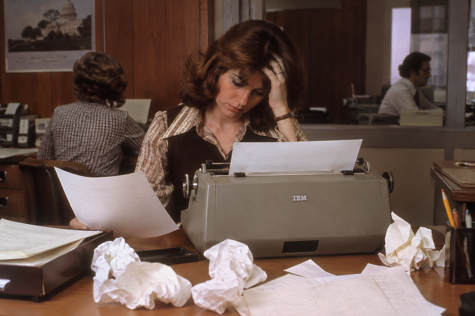 A woman with her hand on her head types on an IBM Selectric typewriter at a desk cluttered with papers and crumpled balls of paper.