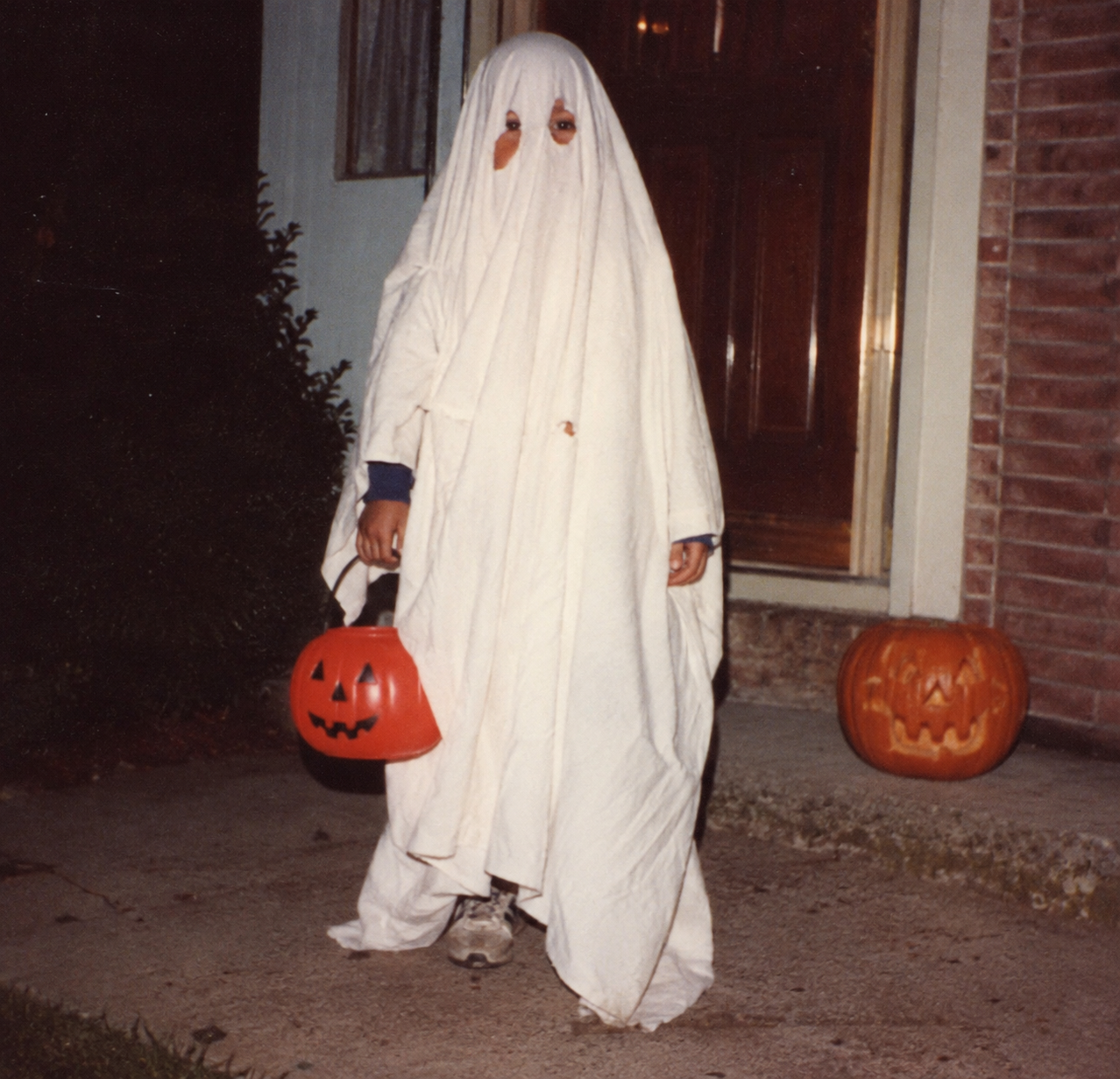A child dressed in a ghost costume made from a white sheet, holding an orange plastic pumpkin bucket, standing in front of a house next to a carved jack-o’-lantern. 1980s Halloween costumes.