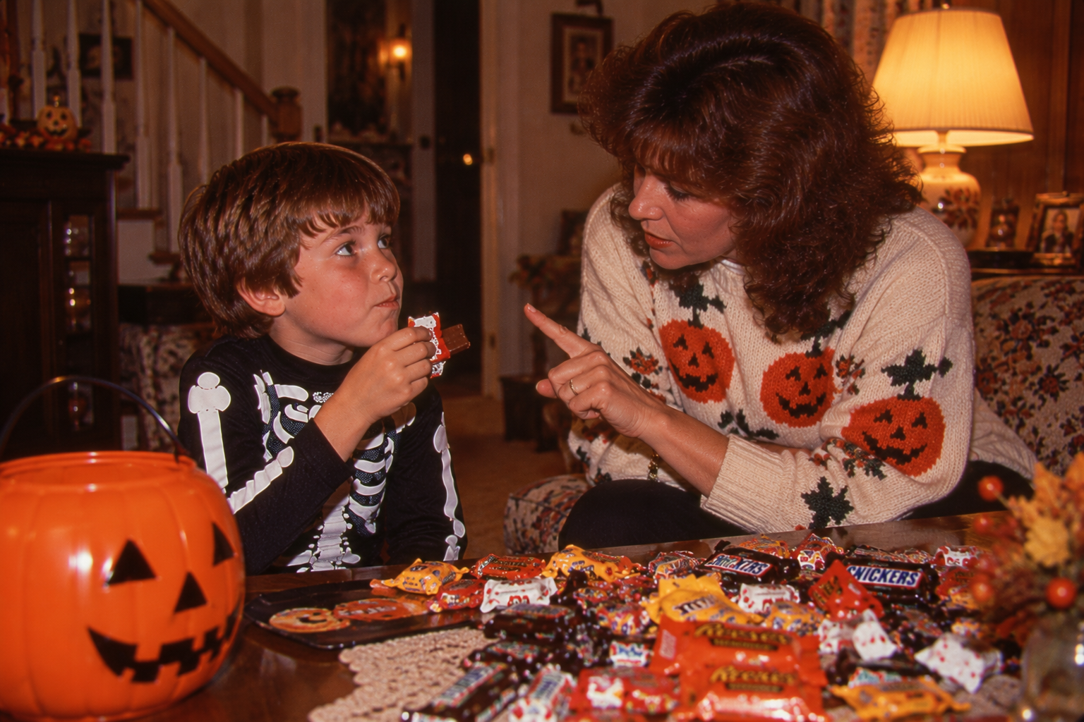 A woman in a pumpkin sweater talks to a boy in a skeleton costume as they look through a pile of Halloween candy next to a jack-o’-lantern bucket.