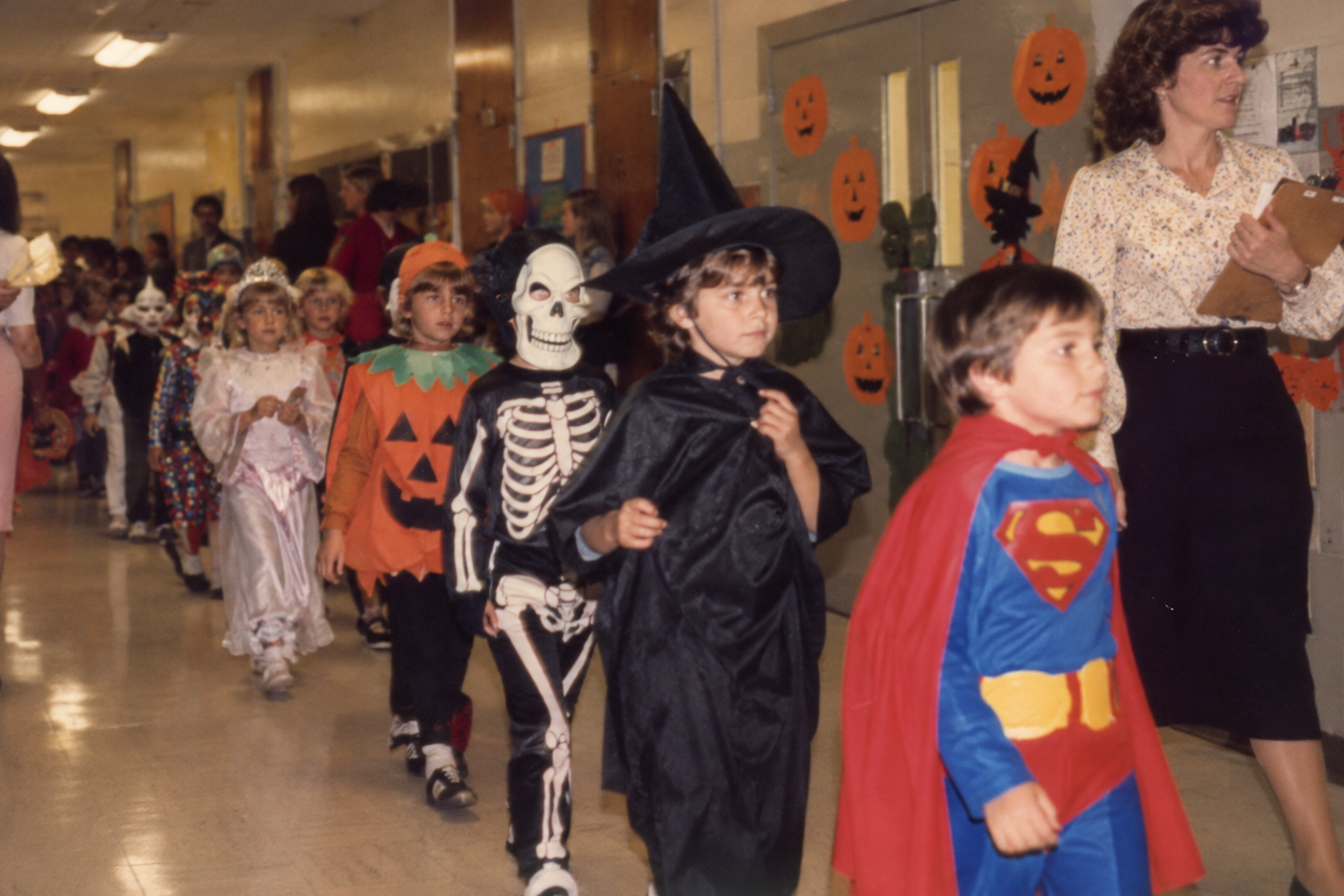 A line of elementary school children in Halloween costumes walking through a school hallway, followed by an adult woman.