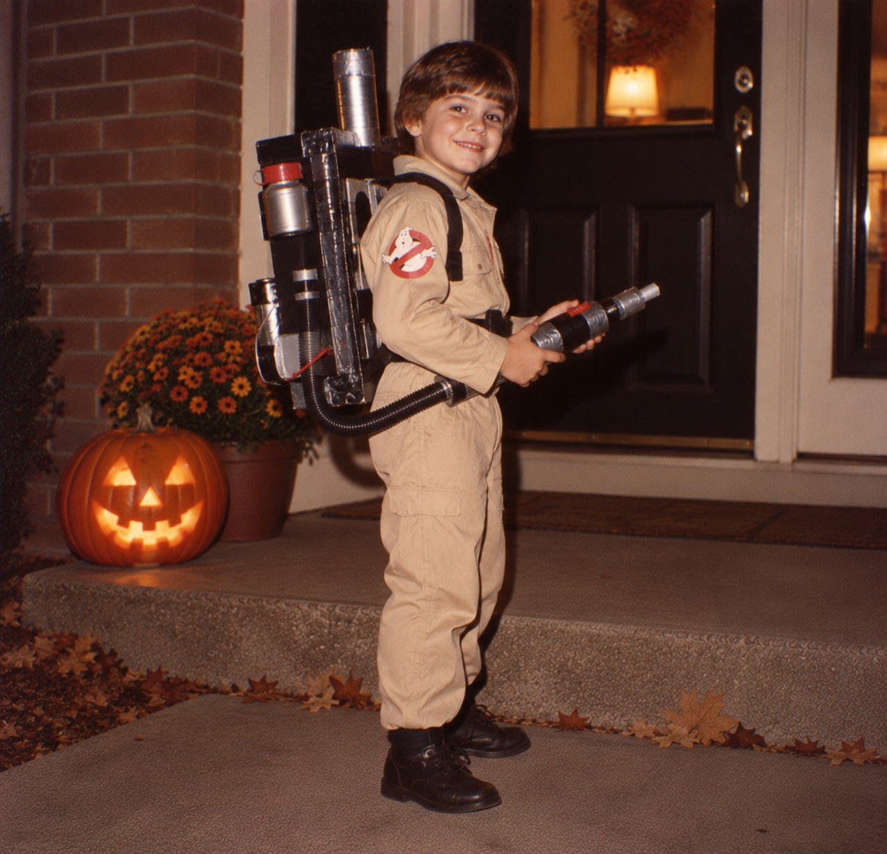 A young boy dressed as a Ghostbuster with a proton pack and neutron wand stands on a porch next to a lit jack-o’-lantern.
