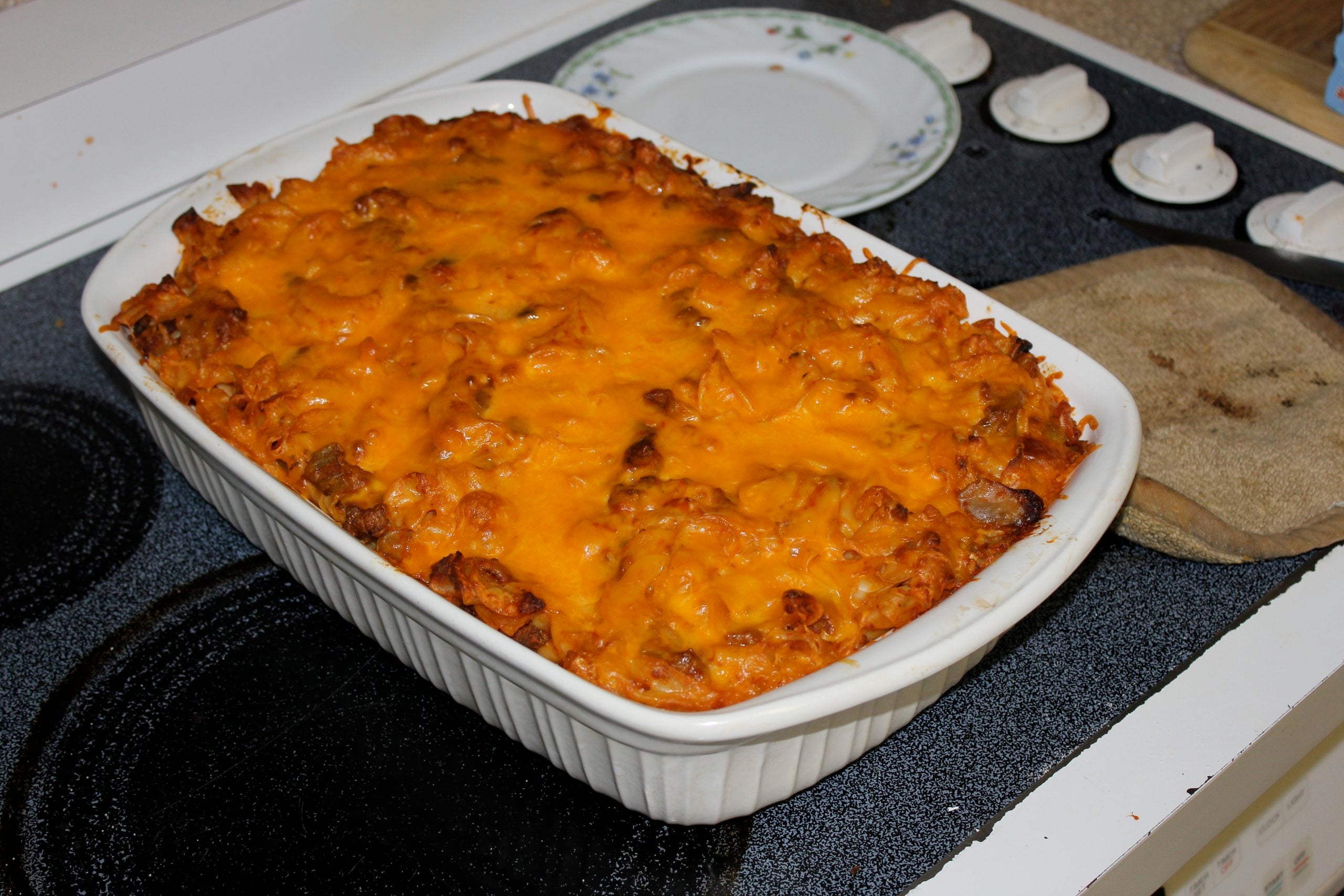 A rectangular white baking dish filled with pasta topped with melted, browned cheddar cheese sitting on a stovetop.