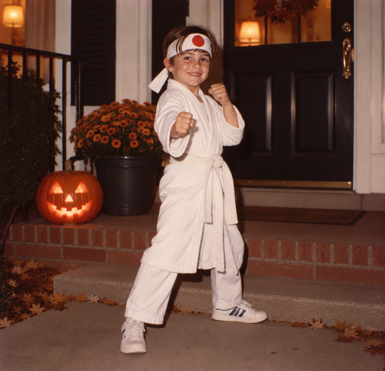 A child wearing a white karate uniform and a headband with a red circle stands on a porch next to a lit jack-o’-lantern.