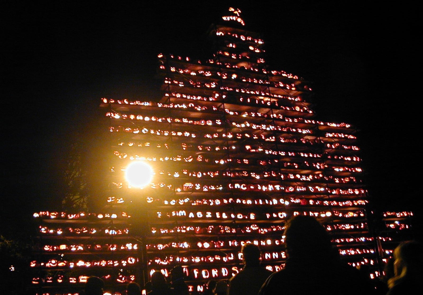 A large, tiered structure stacked with hundreds of glowing, carved jack-o’-lanterns at night.