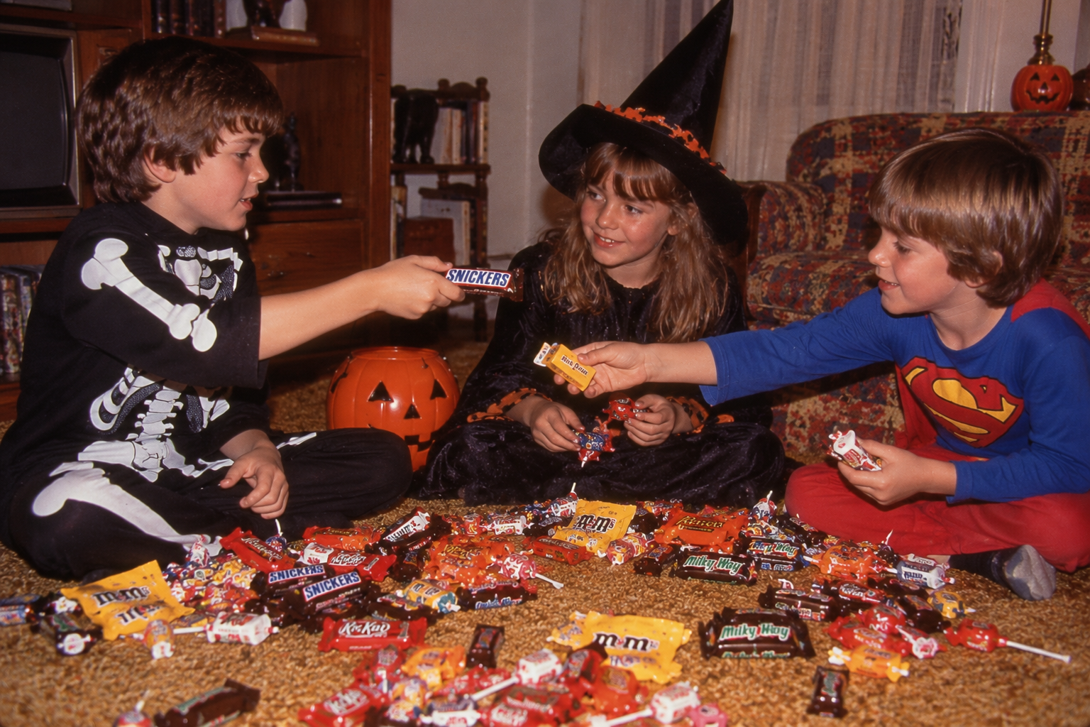 Three children in Halloween costumes sit on a carpet, sharing and sorting a large pile of candy.