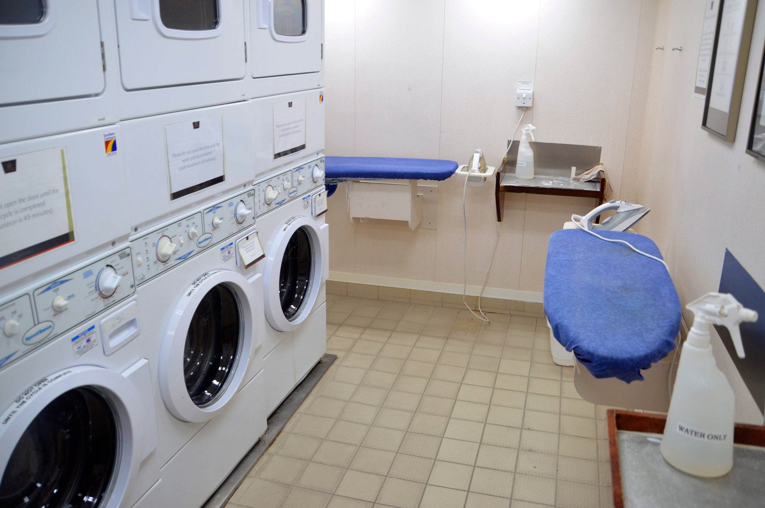 A laundromat room with stacked washers and dryers on the left, two blue ironing boards on the right, an iron, a spray bottle labeled “water only,” and a sink on the back wall.