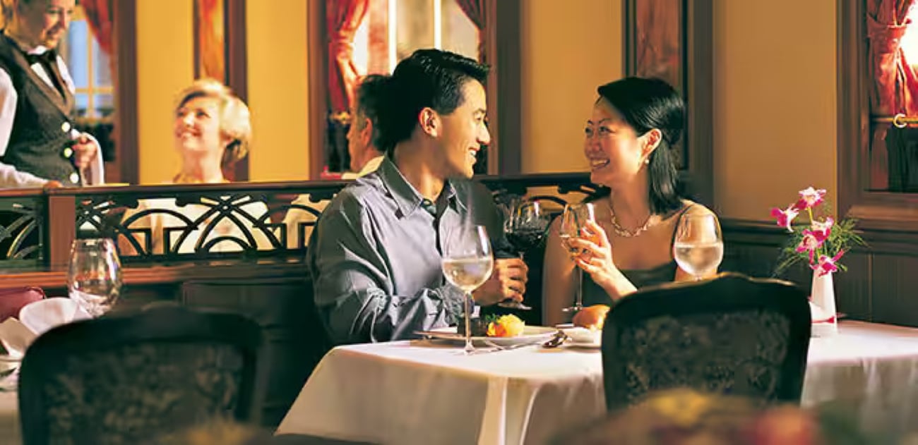 A couple smiling at each other while dining at a restaurant table with wine glasses, as a server stands in the background.