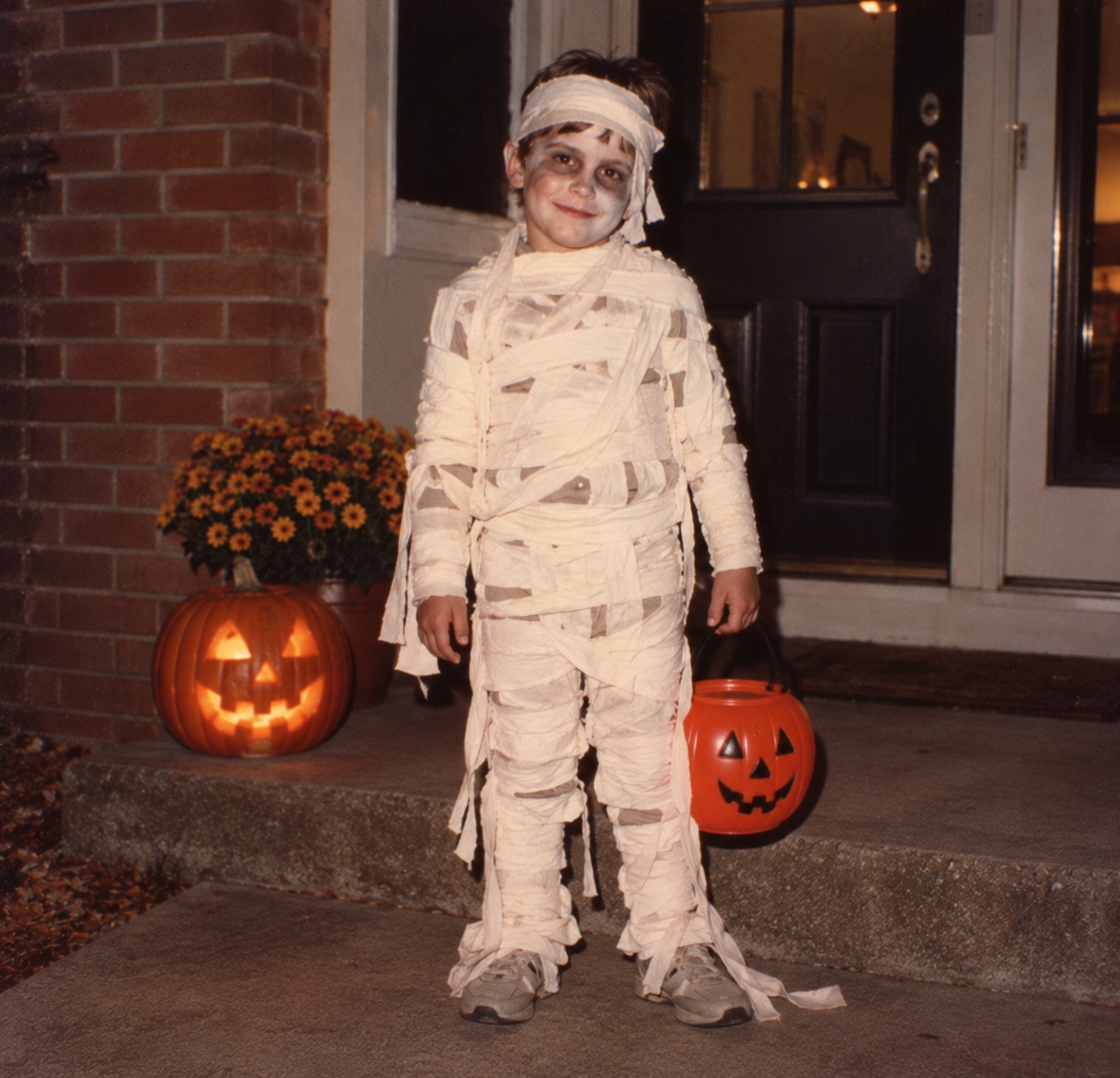 A child in a mummy costume stands on a porch next to a glowing jack-o’-lantern, holding a plastic pumpkin candy bucket.