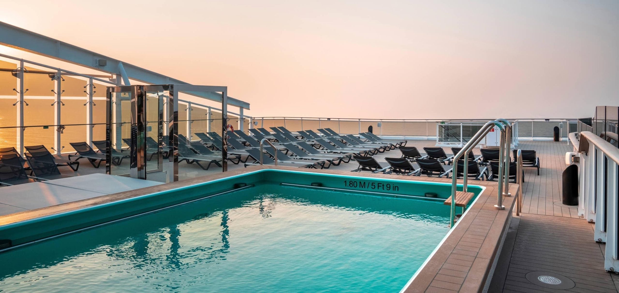 A cruise ship deck featuring a swimming pool with a depth marker of 1.80 M / 5 Ft 9 in, surrounded by rows of empty lounge chairs at sunset.
