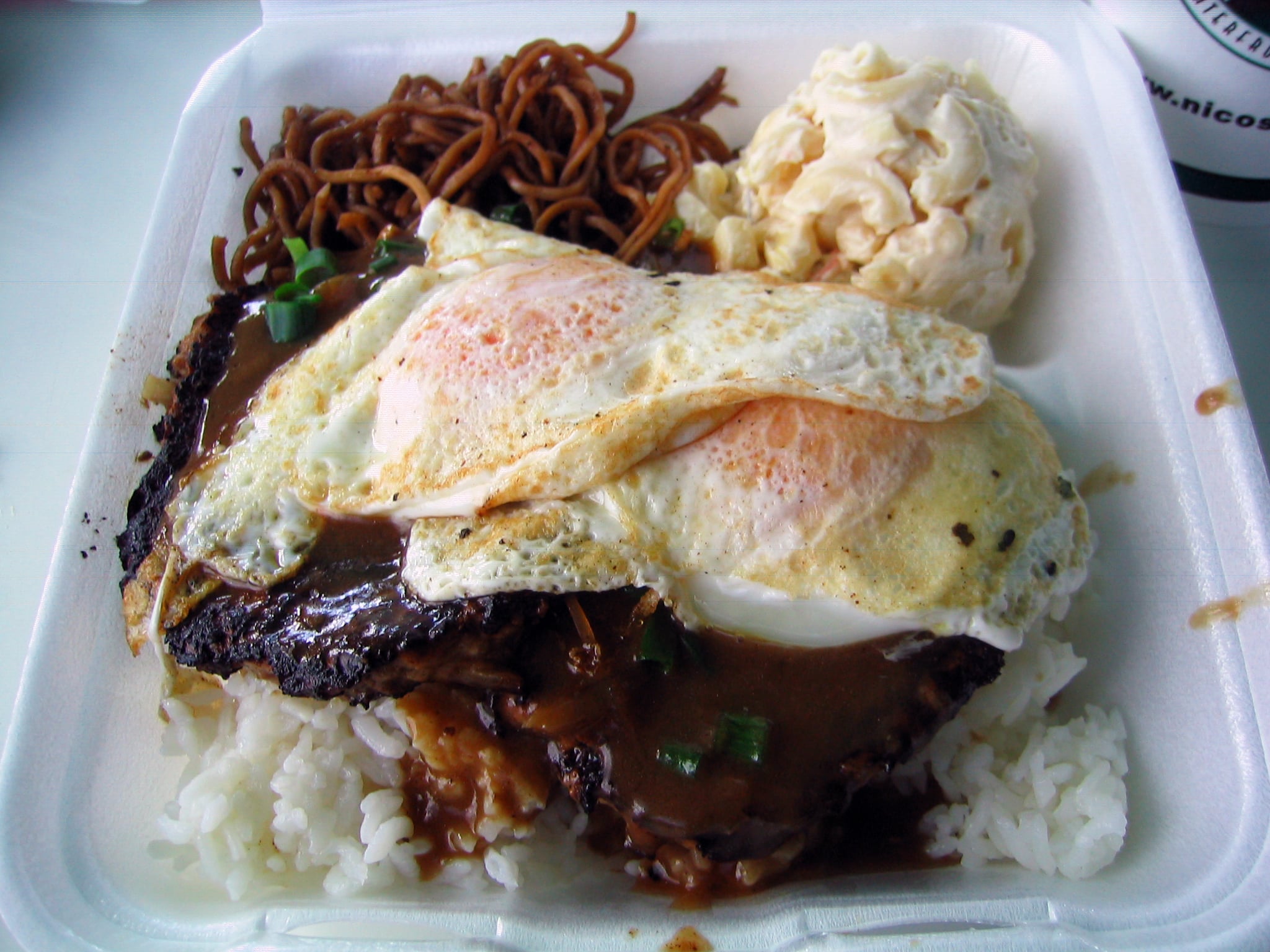 Loco moco served in a styrofoam container with a side of macaroni salad and noodles, reflecting American dining traditions.