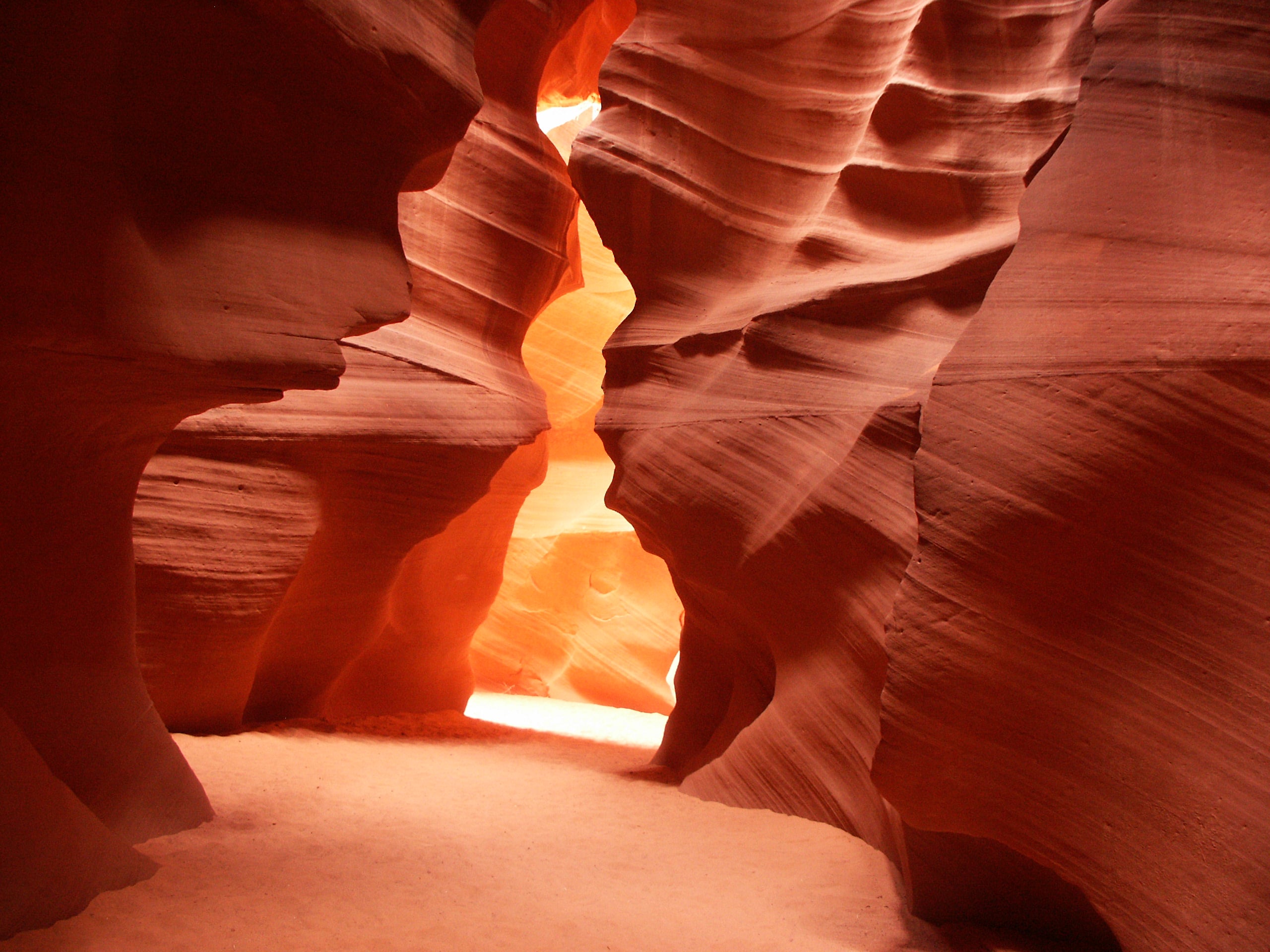 Smooth, undulating red sandstone walls of a narrow slot canyon with a sandy floor.