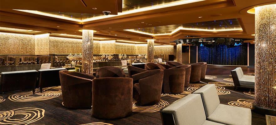 A dimly lit lounge featuring rows of plush armchairs, circular booth seating, and a small stage with blue curtains.