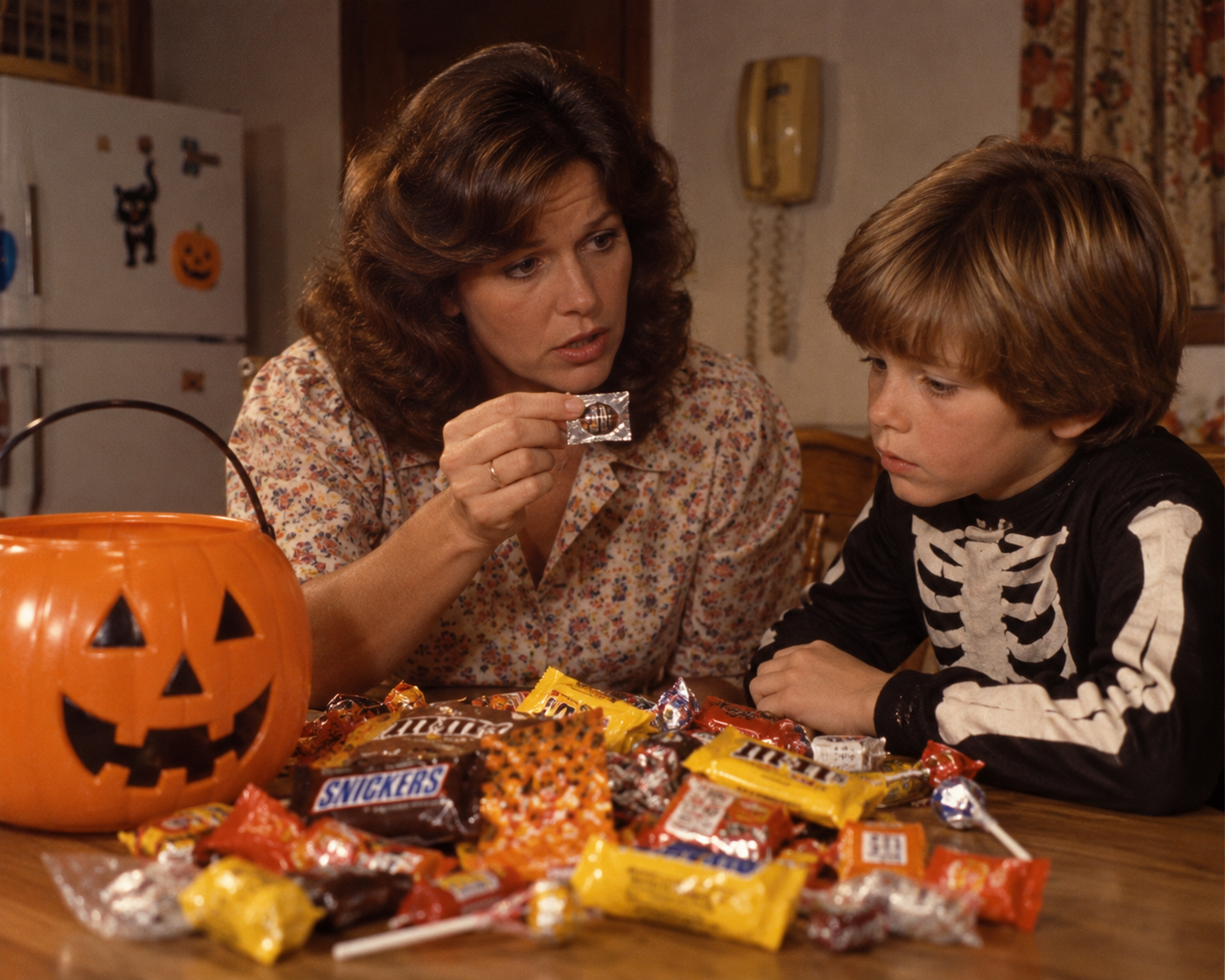 A woman sits with a young boy in a skeleton costume at a table covered in Halloween candy, holding up a small treat for him to see next to a plastic jack-o’-lantern bucket.