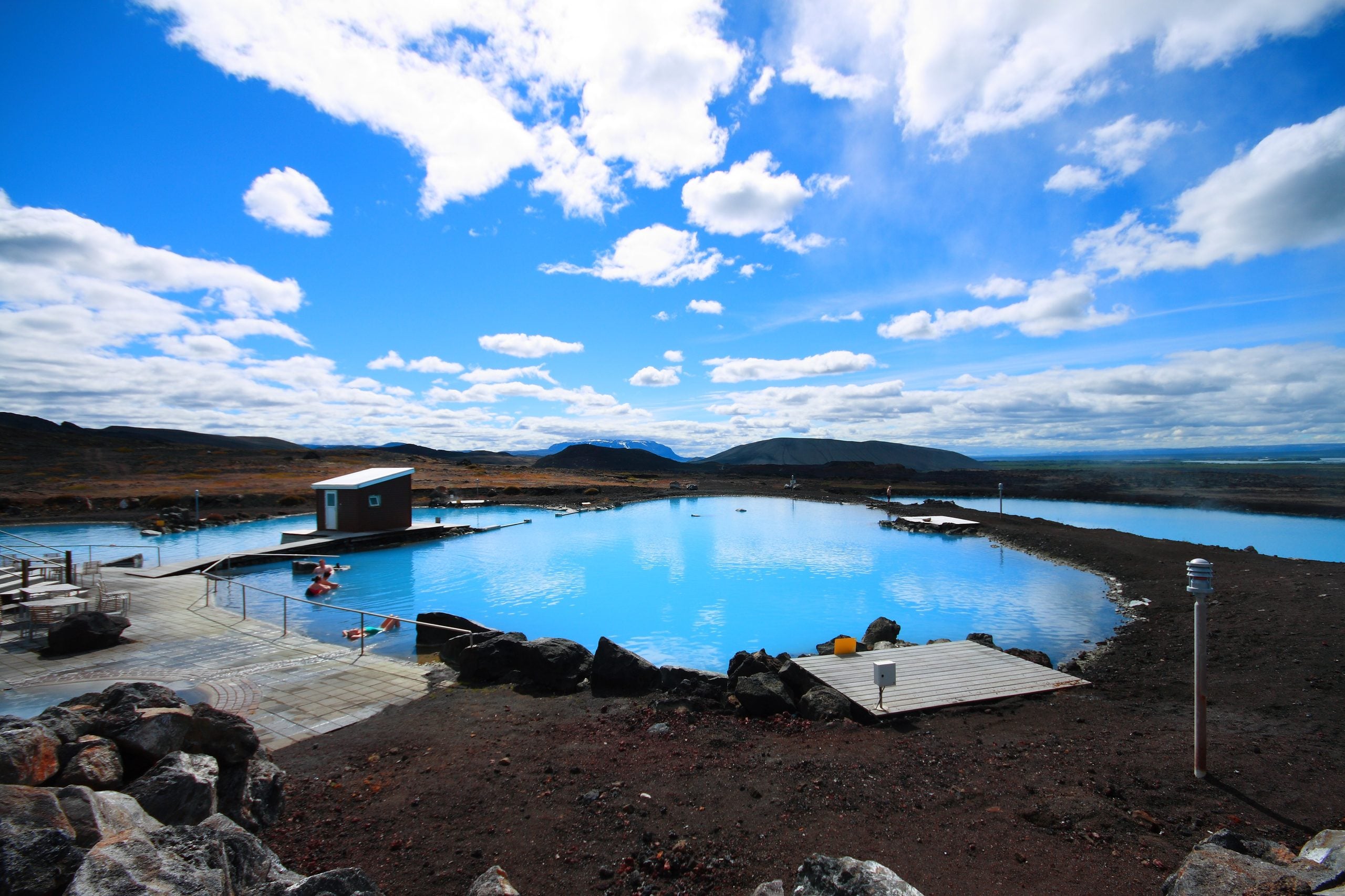 People soak in a bright blue geothermal pool surrounded by dark volcanic rock under a partly cloudy sky.