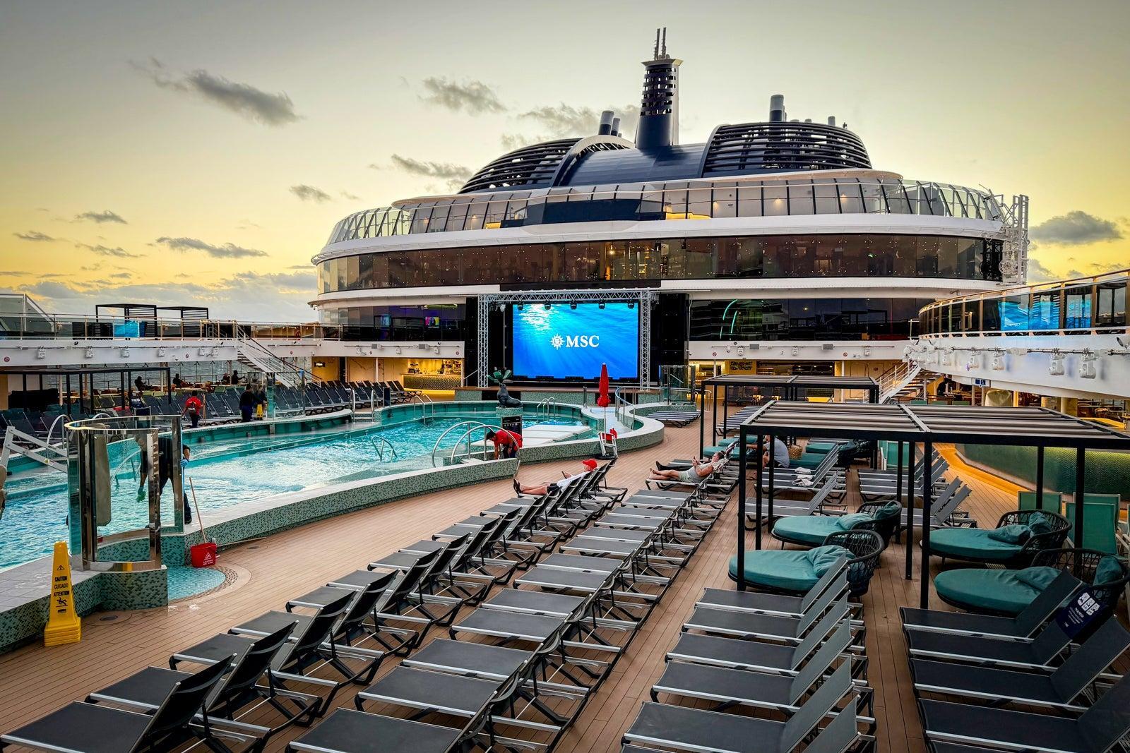 A cruise ship deck featuring a swimming pool, rows of lounge chairs, and a large outdoor screen displaying the MSC logo at sunset.