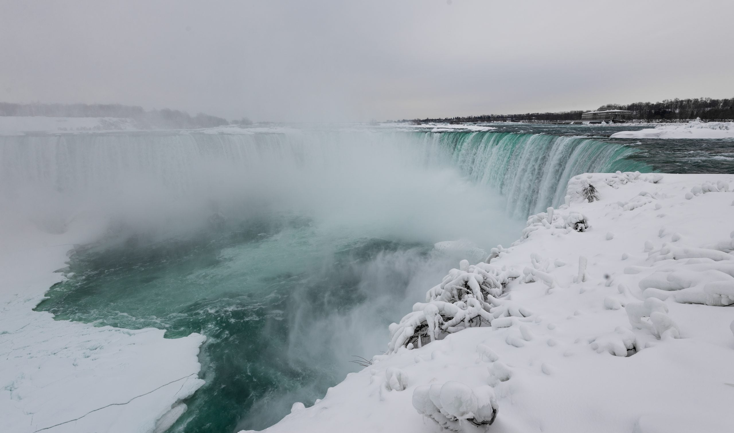 Niagara Falls in winter with snow-covered rocks in the foreground and mist rising from the turquoise water, ideal for trips where bad weather creates a stunning icy landscape.
