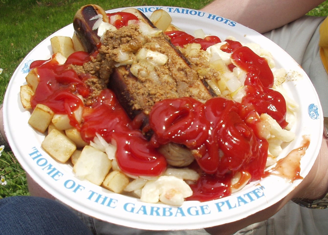 A Nick Tahou Hots garbage plate on a paper plate, featuring a hot dog topped with meat sauce and onions, served over home fries and macaroni salad, all covered in ketchup.