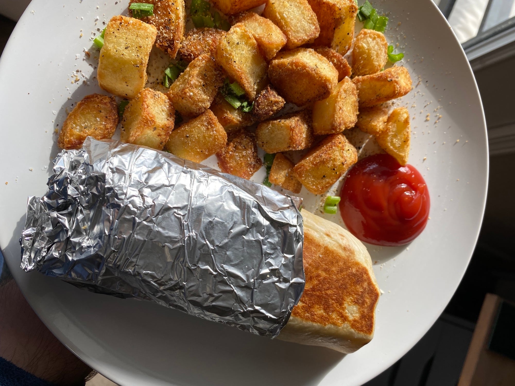 A burrito partially wrapped in foil, served on a plate with crispy diced potatoes and a side of ketchup.