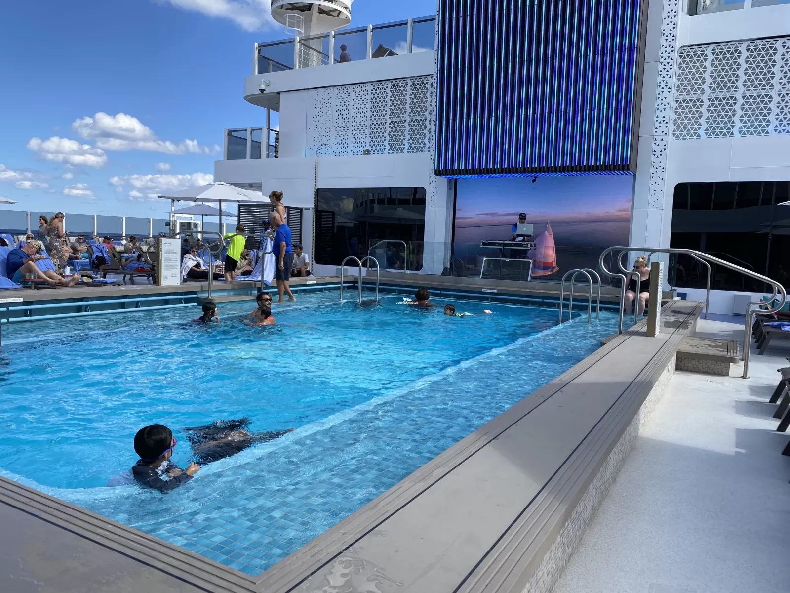 People swimming in a cruise ship pool with a DJ performing on a stage in the background.