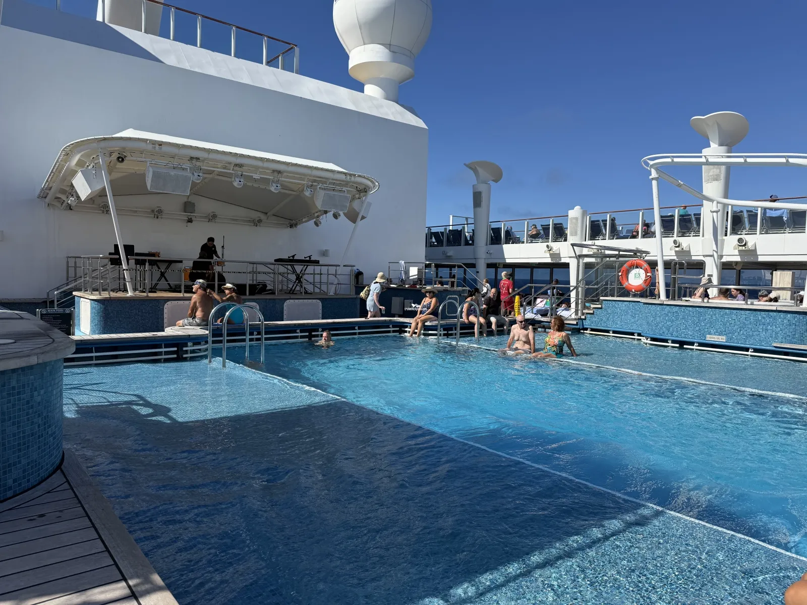 People relaxing in and around a cruise ship pool while a musician performs on a stage nearby.