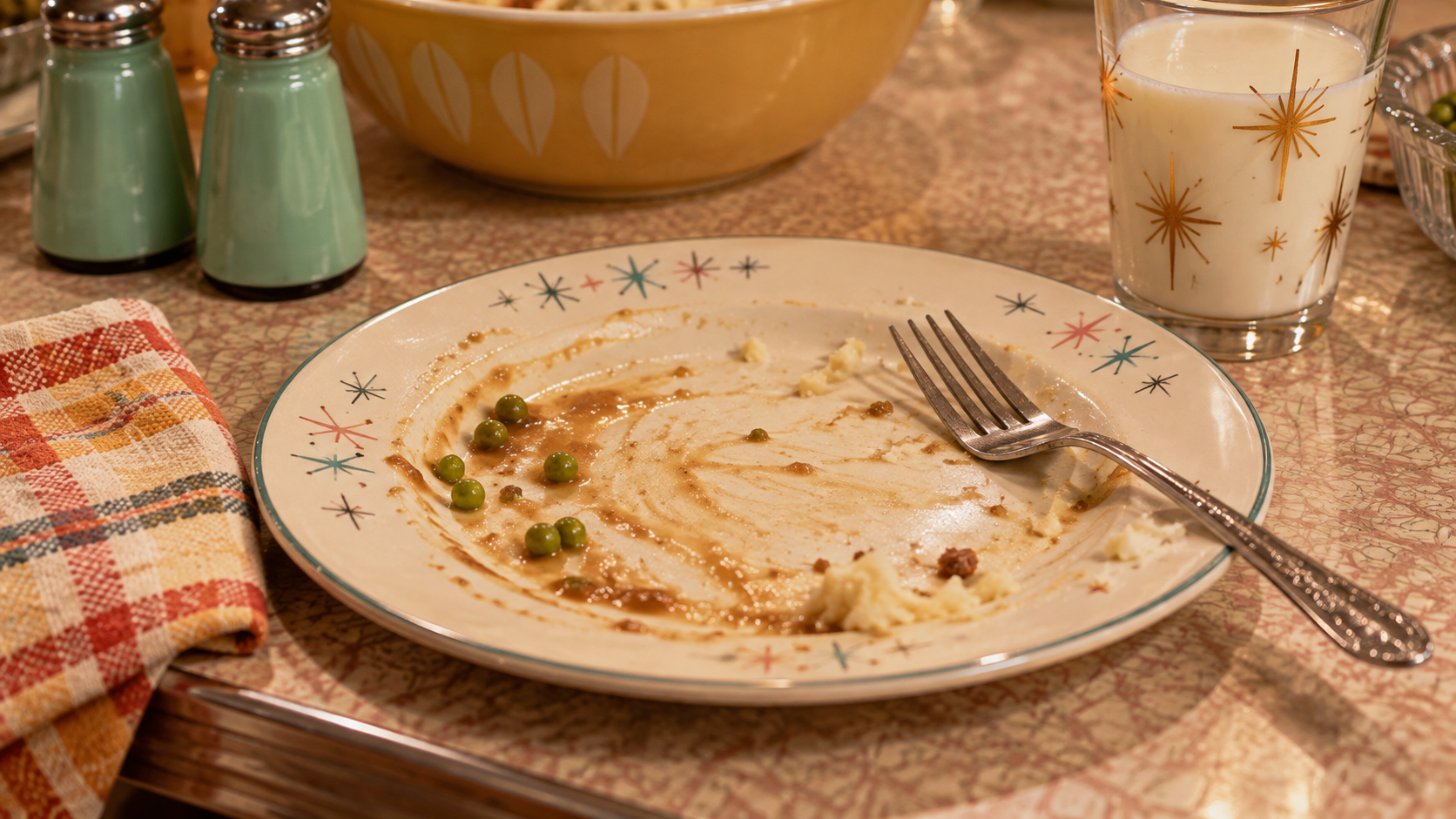 A dinner plate with leftover peas and gravy, a fork, a plaid napkin, and a glass of milk on a patterned table.
