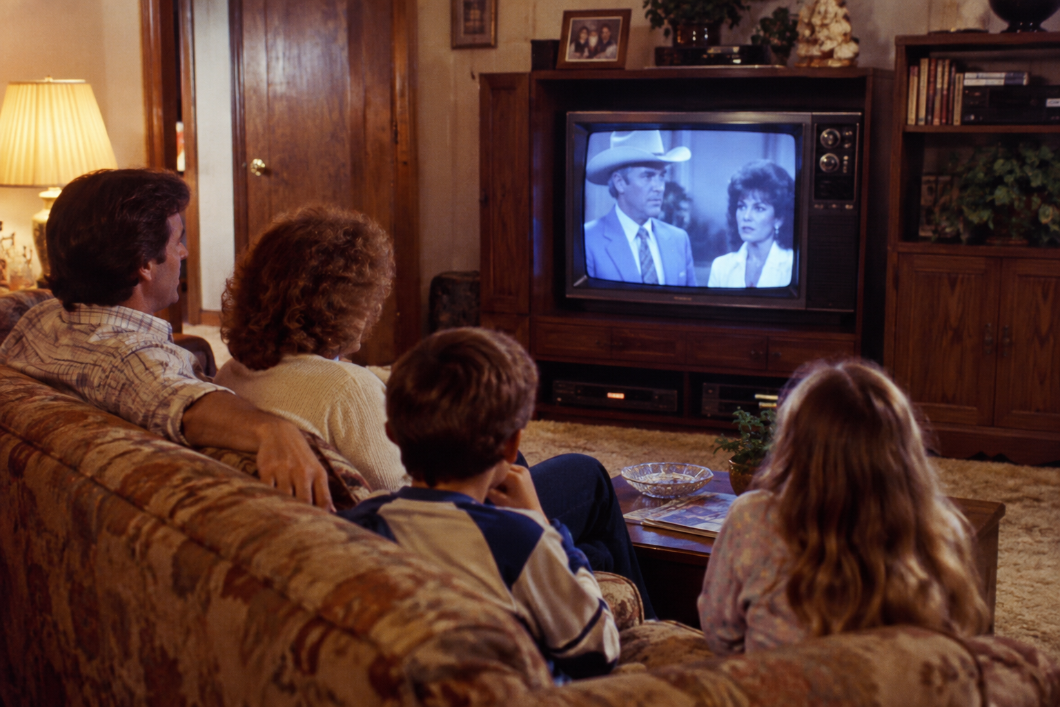 A family sits on a sofa watching a television show featuring a man in a cowboy hat and a woman in a suit. Friday nights in the 1980s.