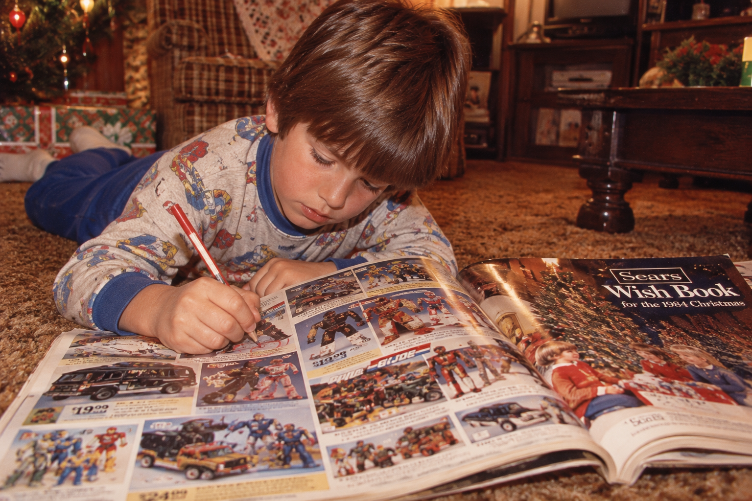 A young boy lying on a carpeted floor uses a red pen to circle toys in a 1984 Sears Wish Book.