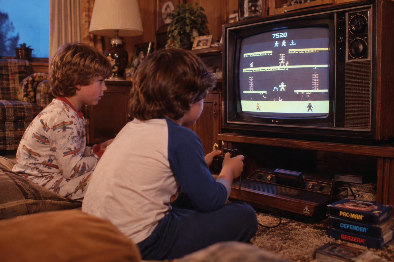 Two children in pajamas sit on the floor playing a retro video game on an old TV. Game cartridges and boxes, including Pac-Man and Defender, are visible on the carpet nearby. The room has cozy, vintage decor.