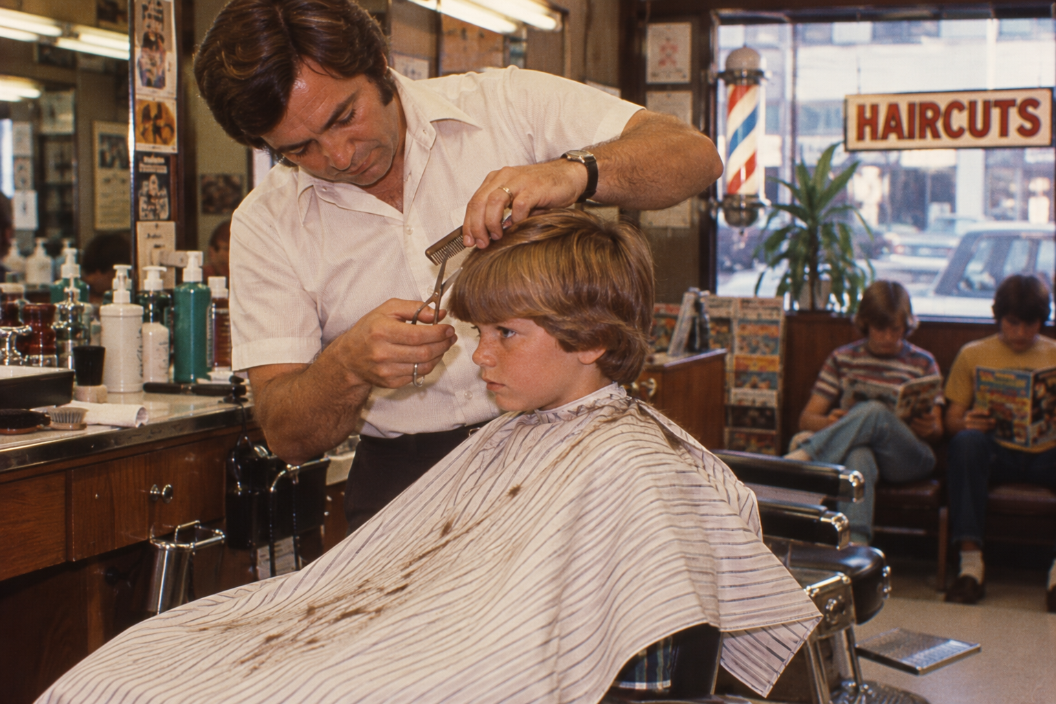 A barber in a light-colored shirt uses scissors and a comb to cut a young boy’s hair in a barbershop.