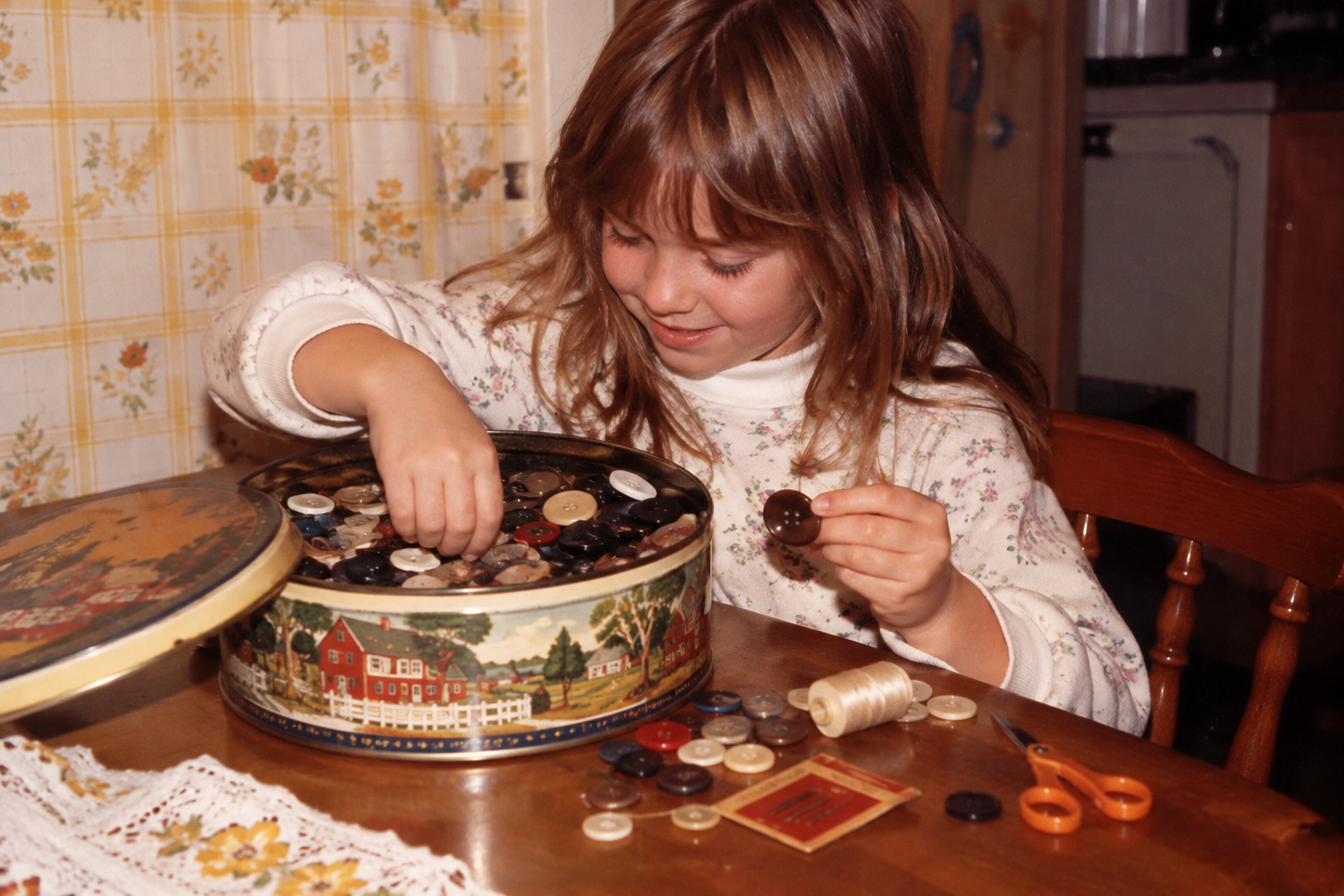 A young girl sits at a table sorting through a round tin filled with various buttons. Grandma's house in the '80s.