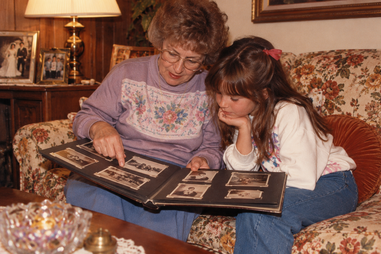 An older woman and a young girl sit on a floral sofa looking at a photo album together.