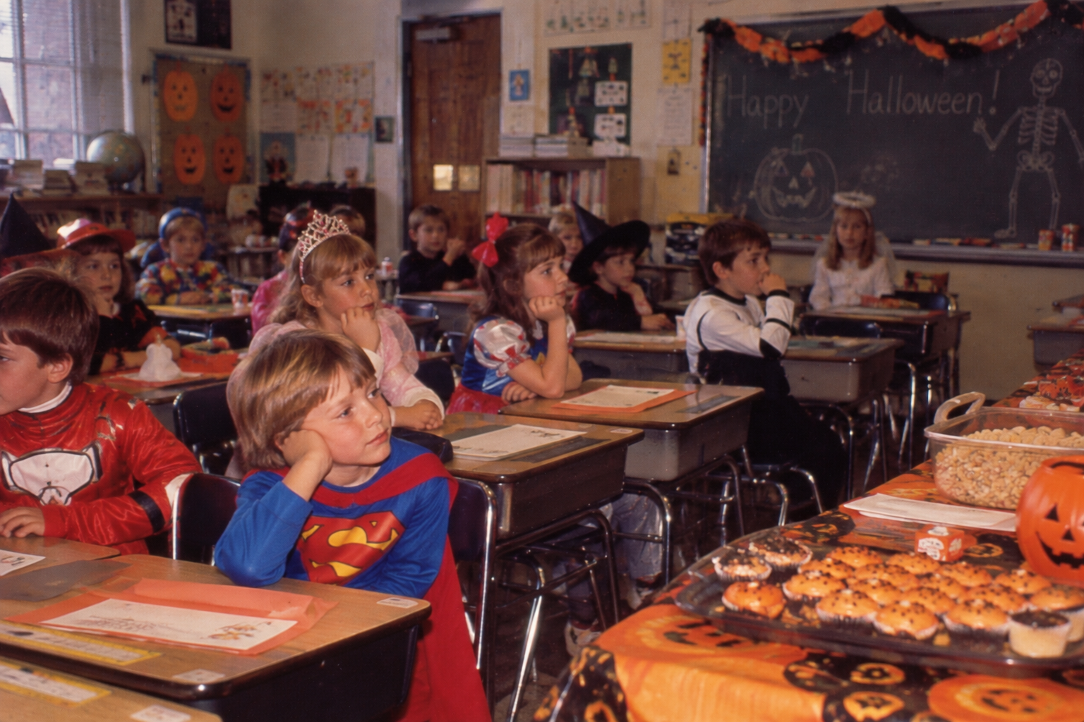 Elementary students in Halloween costumes sit at their desks in a decorated classroom with treats on a table in the foreground.