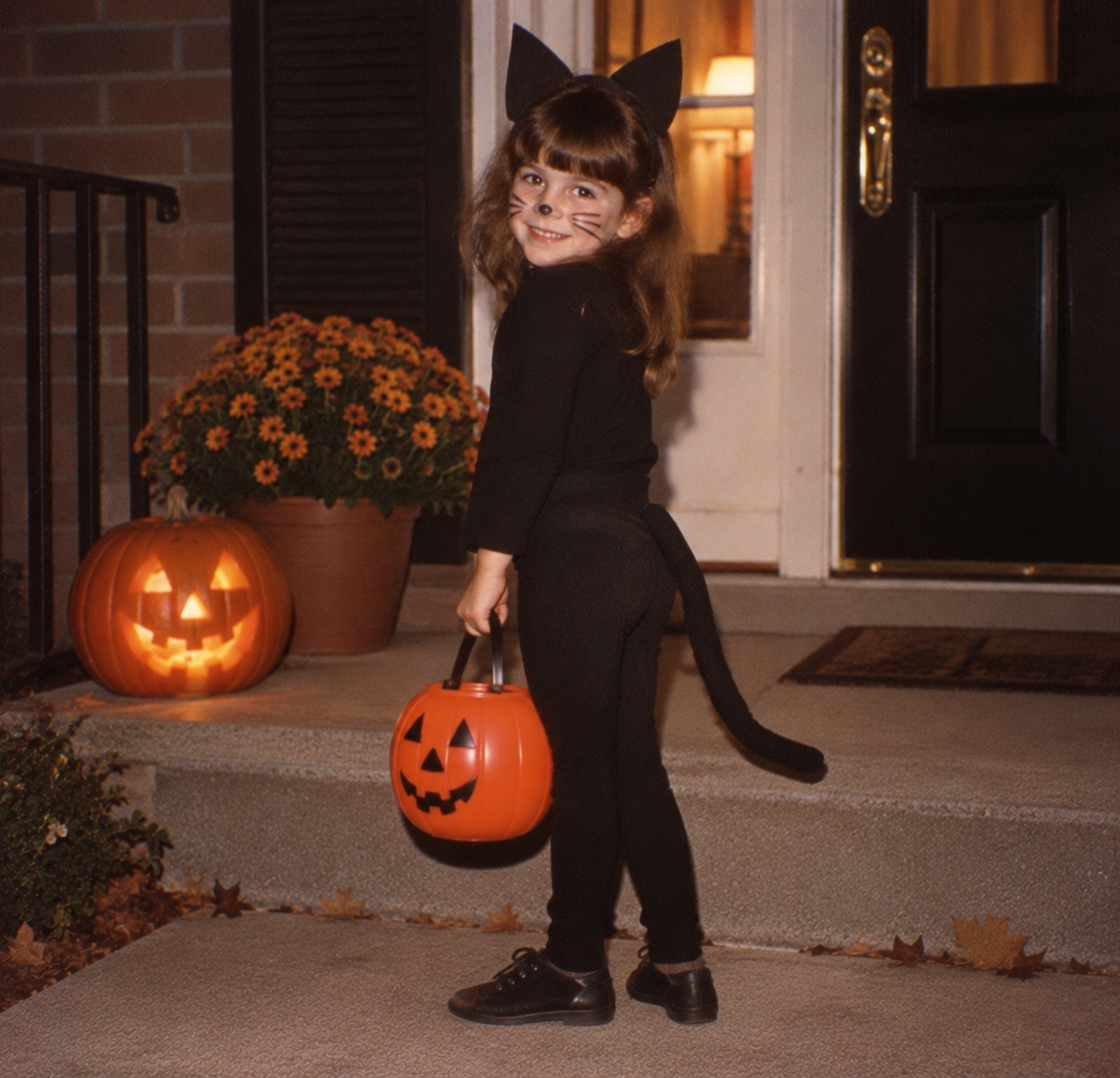 A young girl in a black cat costume with ears and a tail holding a plastic jack-o’-lantern bucket on a porch next to a lit carved pumpkin.