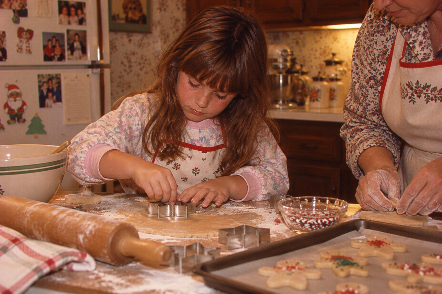 A young girl uses a cookie cutter to cut shapes from dough on a floured surface while an adult assists nearby.