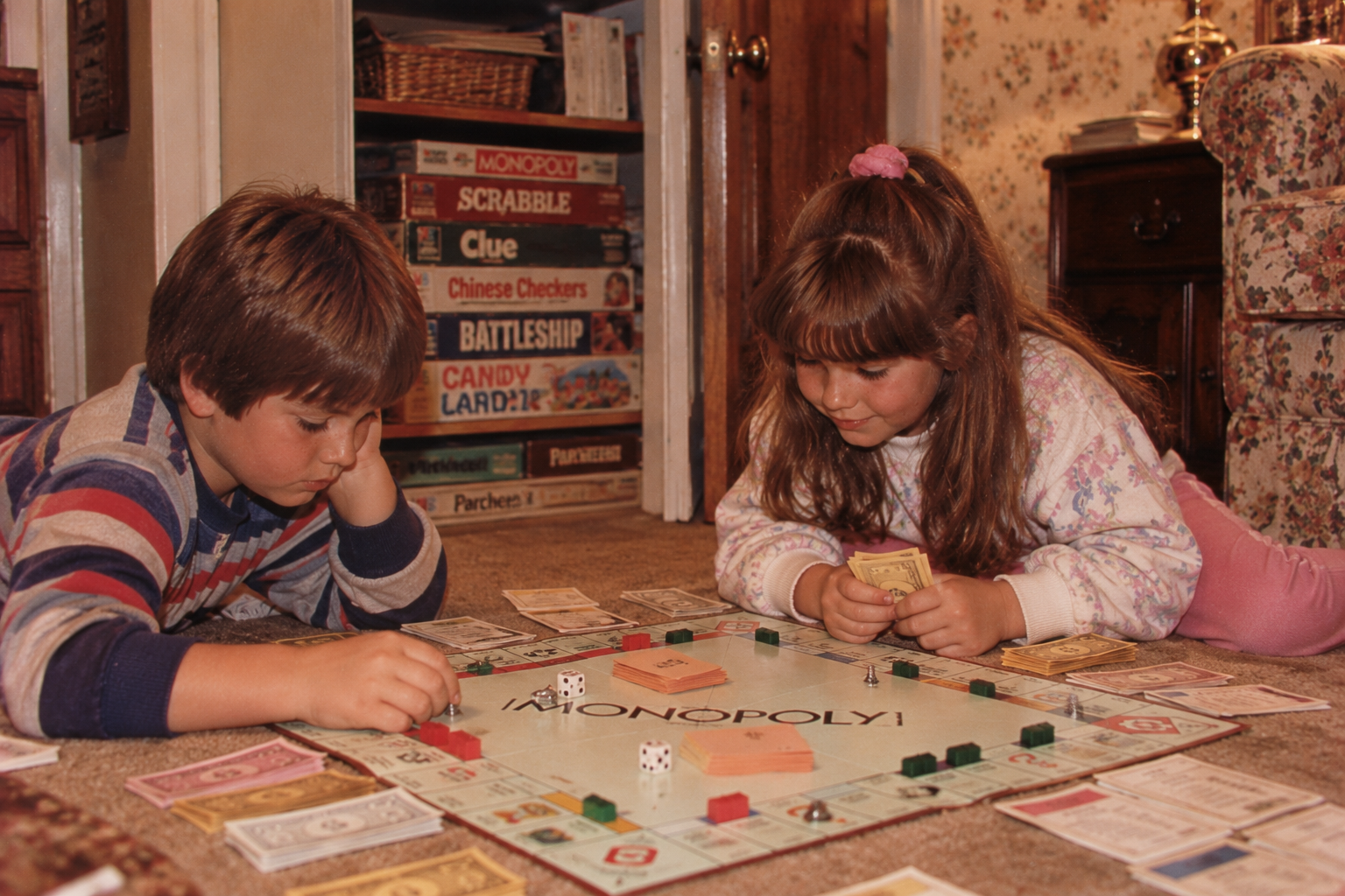 A boy and girl sitting on a carpeted floor playing a game of Monopoly.