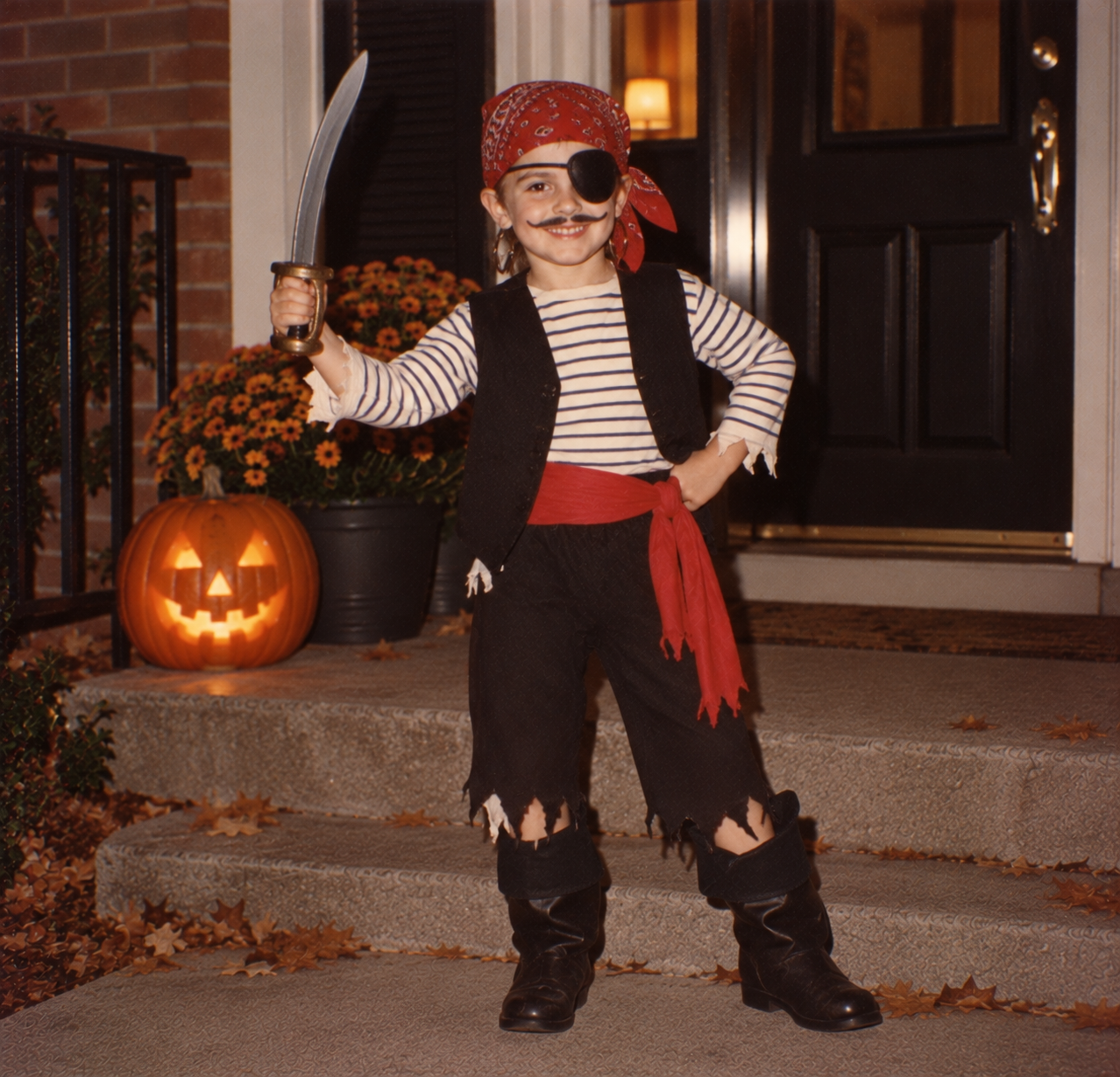 A young child dressed as a pirate with a fake mustache, eyepatch, and plastic sword standing on a porch next to a lit jack-o’-lantern.