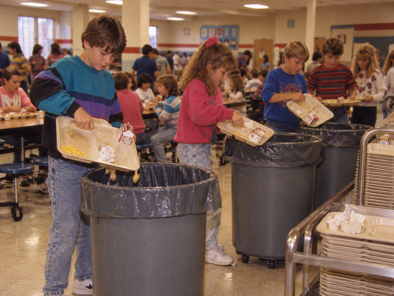 Students in a school cafeteria scrape food from their trays into large trash cans.
