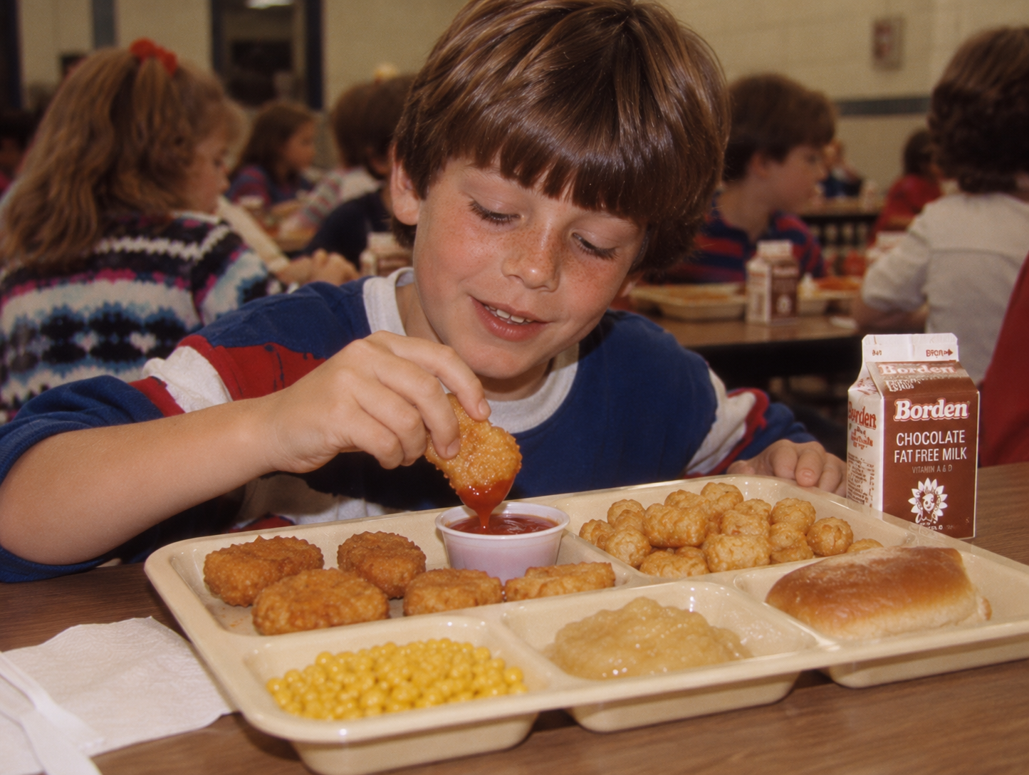 A boy at a school cafeteria table dips a chicken nugget into a small cup of ketchup, with a tray of nuggets, tater tots, corn, applesauce, a dinner roll, and a carton of Borden chocolate milk in front of him.