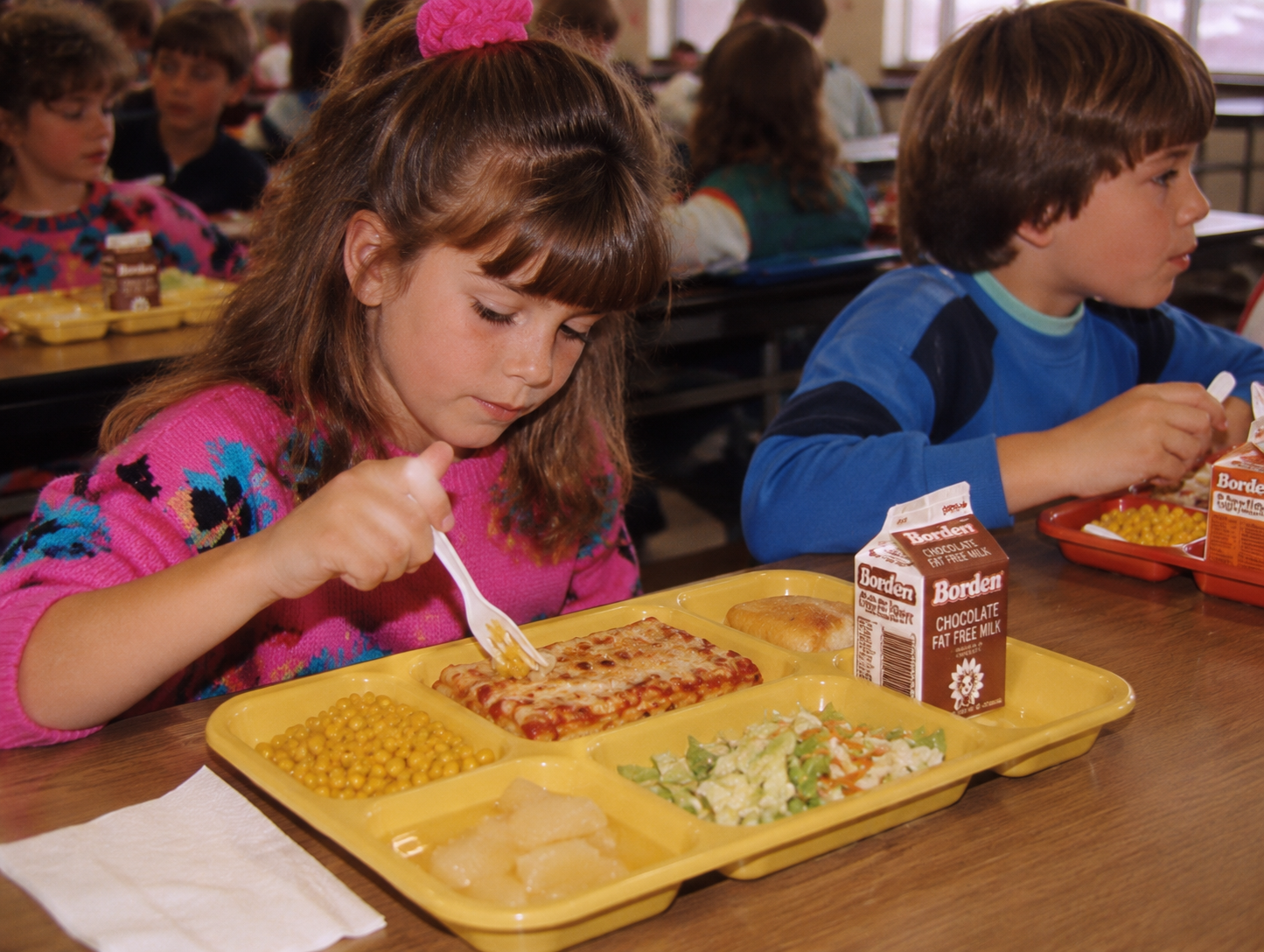 A young girl in a pink sweater eats a school lunch of pizza, corn, coleslaw, and fruit from a divided yellow tray, accompanied by a carton of Borden chocolate milk.