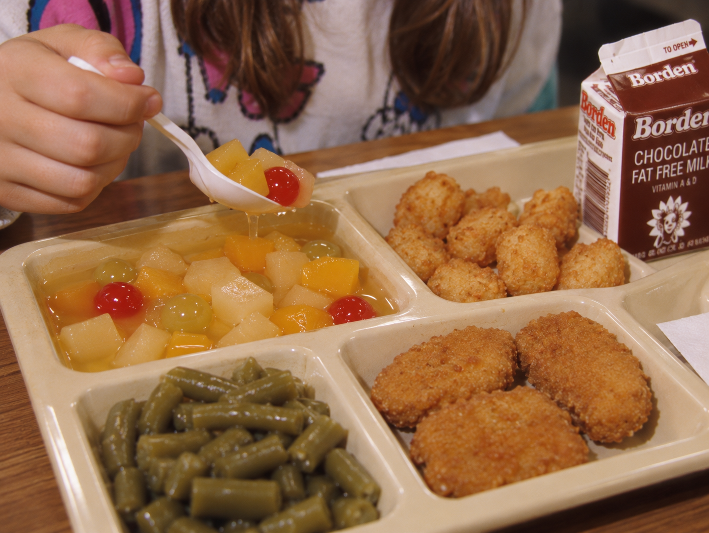 A child uses a plastic spoon to scoop fruit cocktail from a divided lunch tray containing green beans, tater tots, and chicken nuggets, next to a carton of Borden chocolate milk.