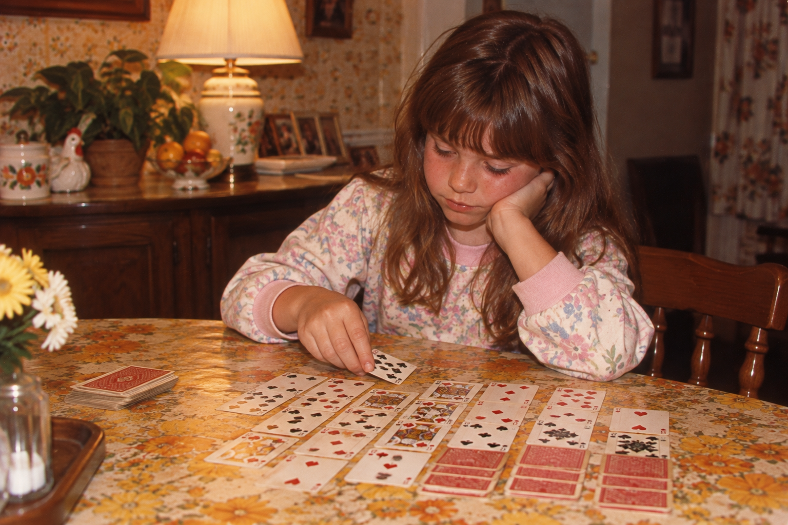 A young girl sits at a table playing solitaire with a deck of cards spread out before her.