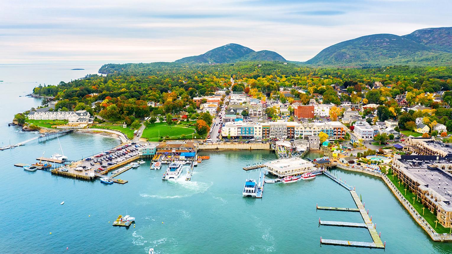 Aerial view of Bar Harbor, Maine, featuring the harbor with docked boats, a town center, and surrounding green mountains under a cloudy sky.
