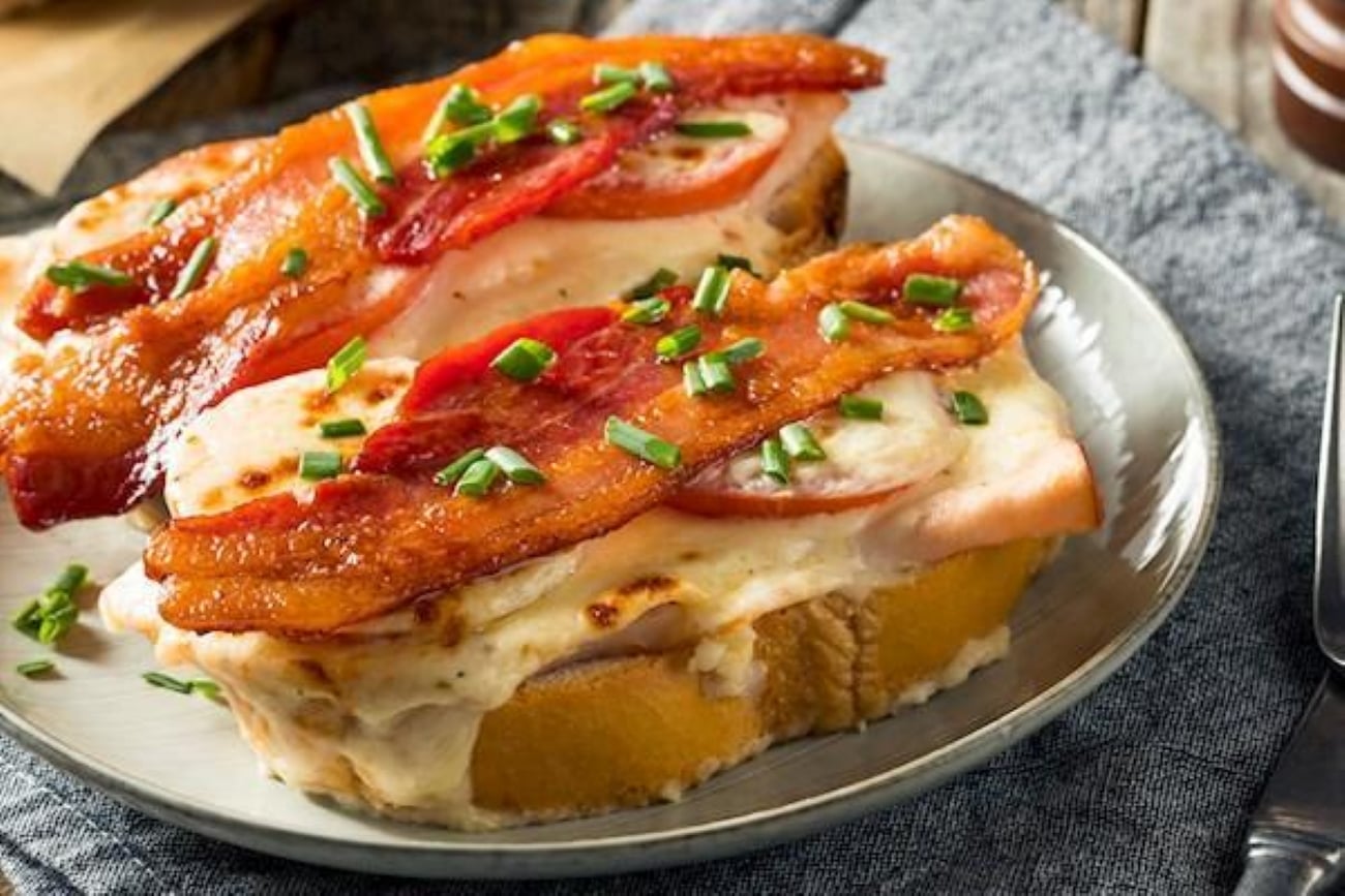Two open-faced sandwiches topped with melted cheese, tomato slices, crispy bacon, and chopped chives on a plate.