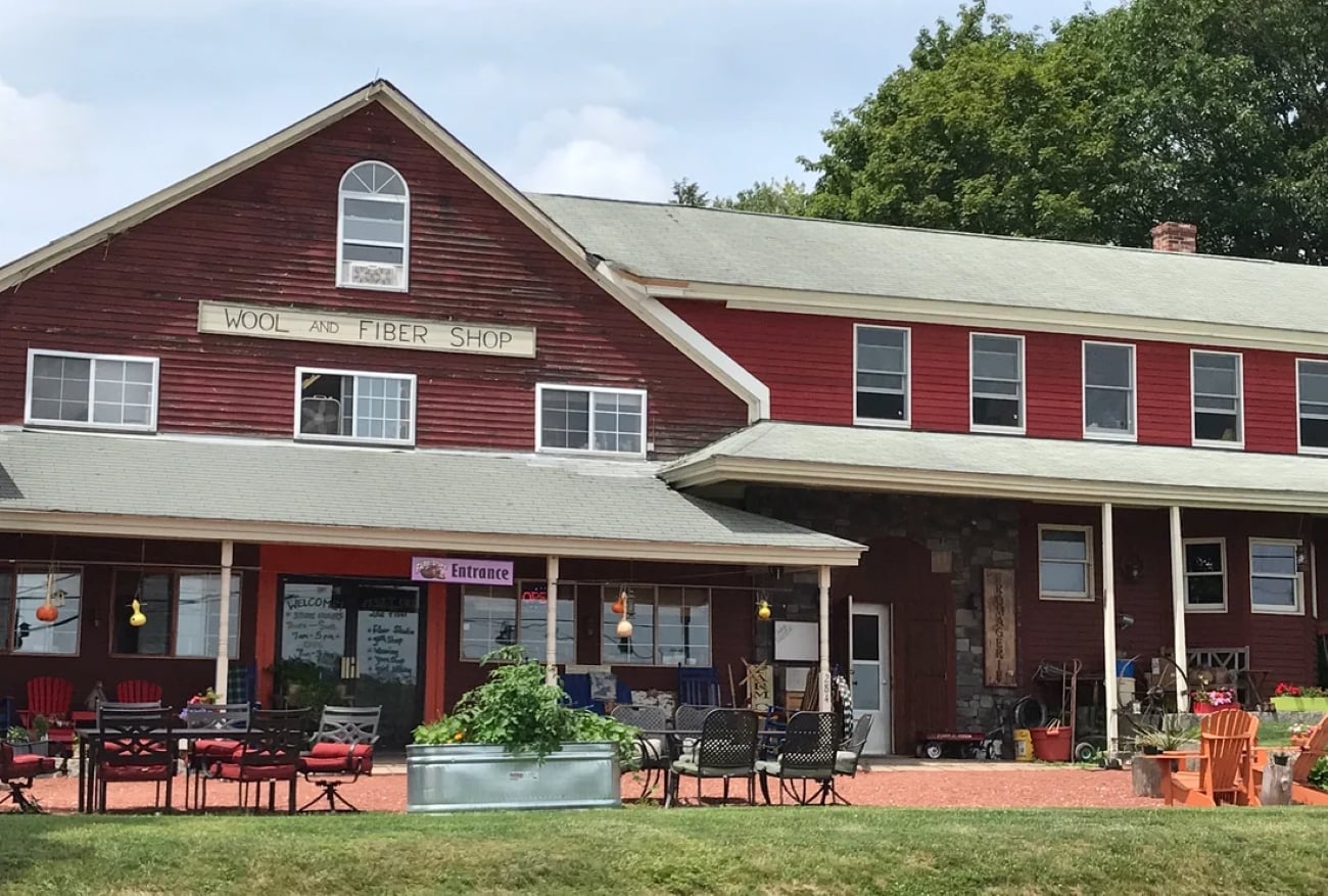 A red barn-style building with a sign that reads “Wool and Fiber Shop” above an entrance with patio furniture on the lawn.
