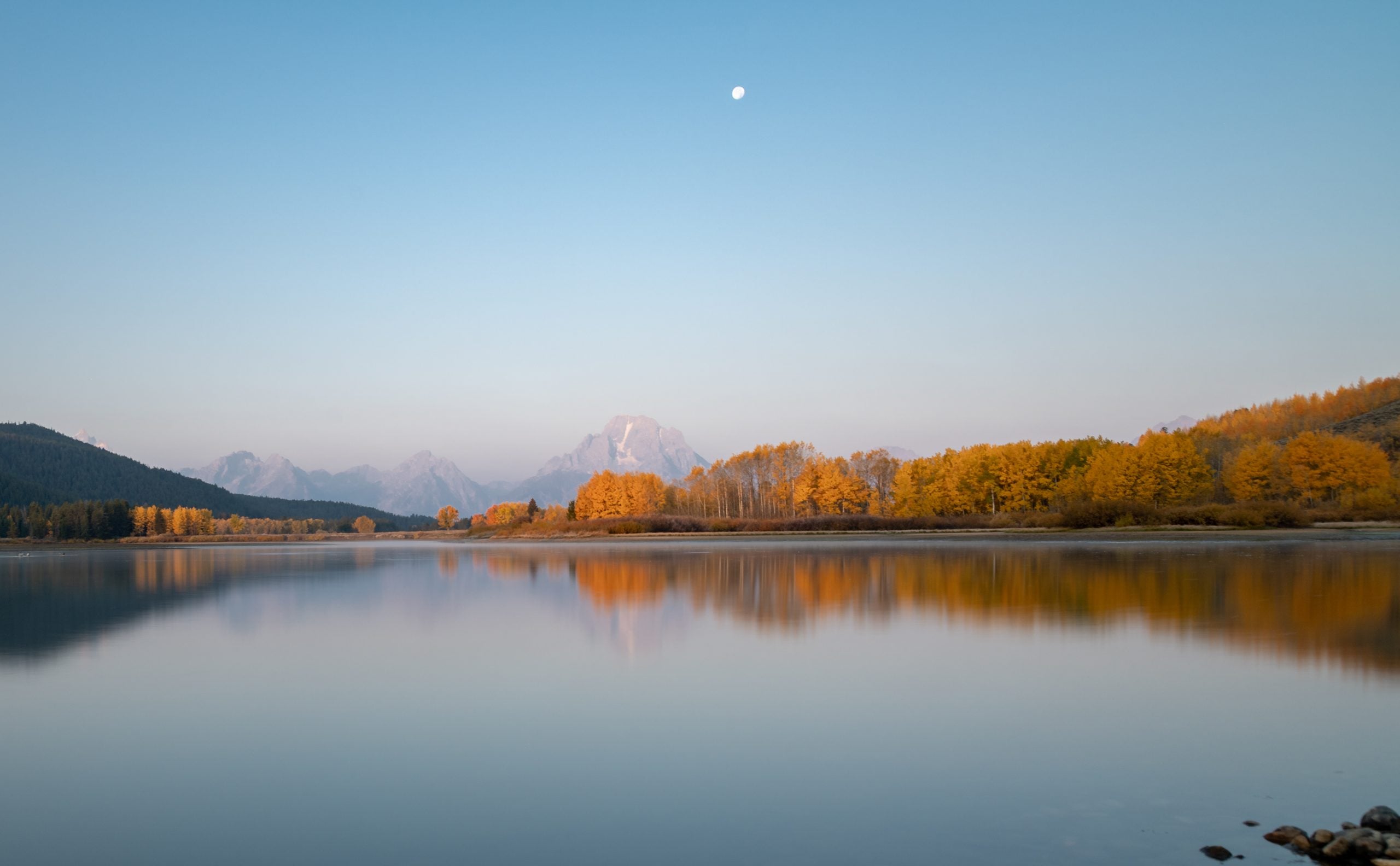 American destinations A calm lake reflects golden autumn trees and snow-capped mountains under a pale blue sky with a small moon.