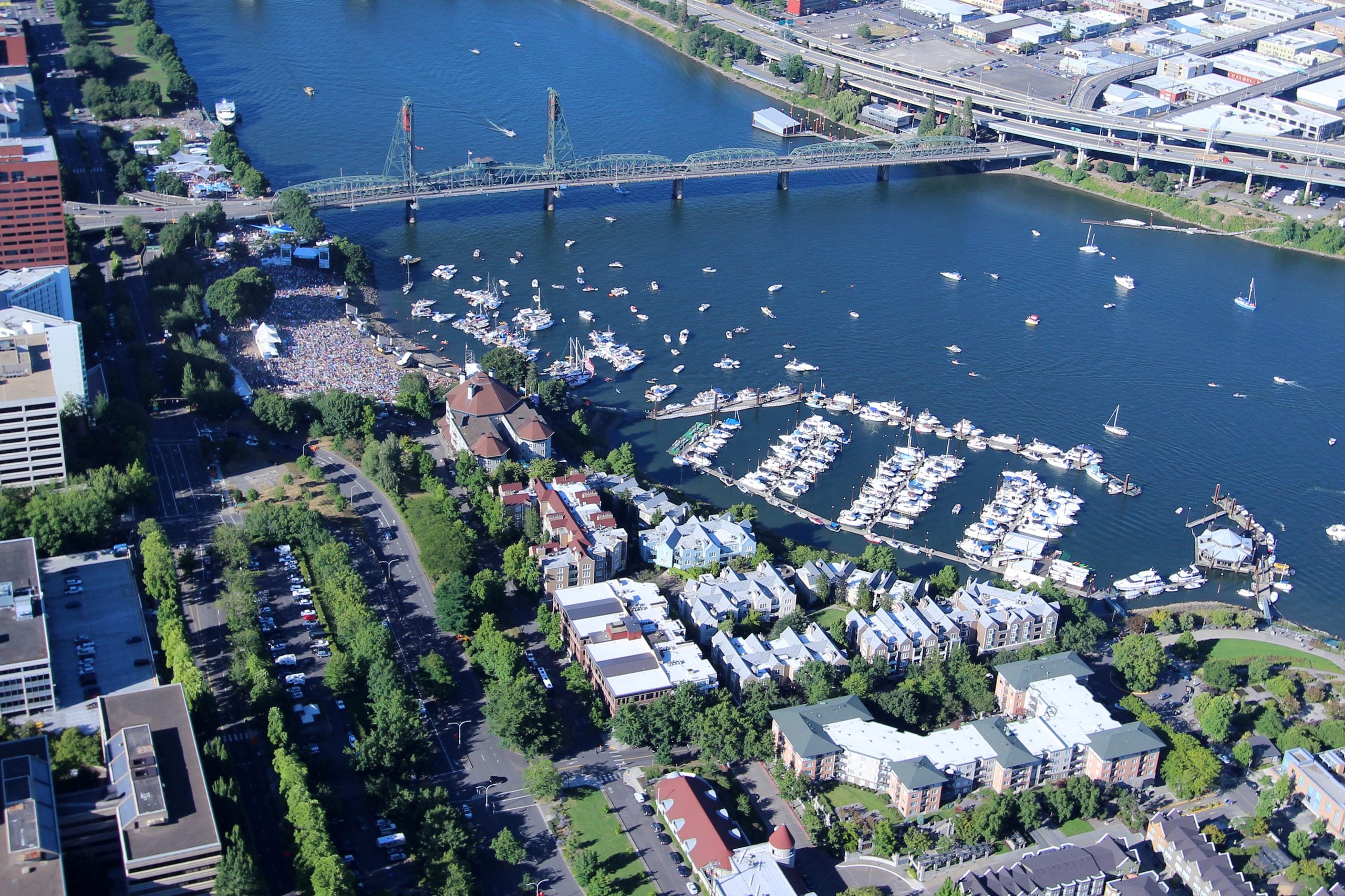 Aerial view of a river with a large bridge, a crowded park hosting an outdoor event, and a marina filled with docked boats.